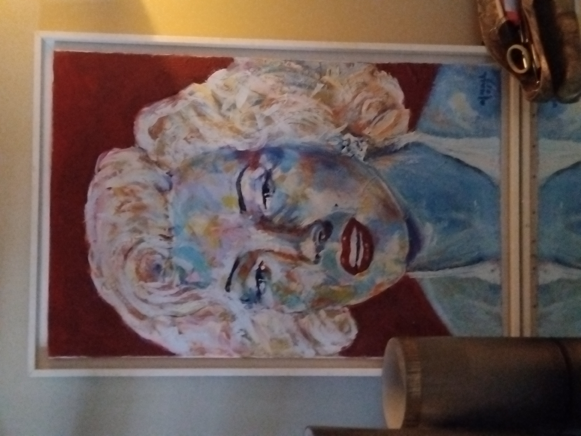 An unframed, vibrant painting of Marilyn Monroe's face, rendered in a bold, abstract style with broad brushstrokes and a multi-colored palette, is displayed prominently. The background of the painting is a deep, muted red. Monroe's signature blonde hair is depicted in swirls of white, yellow, and pink. Her face is a mosaic of blues, greens, pinks, and yellows, with stark black eyebrows and bright red lips. A hint of a white garment is visible beneath her chin. The painting is signed in the lower right corner with a stylized signature and the year "1973". The painting rests on a reflective surface, possibly a glass table, which shows a blurred reflection of the artwork and the objects surrounding it. To the left of the painting, a cylindrical object, possibly a decorative vase or candle holder, made of a textured, dark material stands. To the right of the painting, a small, ornate, metallic dish, likely a trinket tray, holds a ring. The overall lighting suggests an indoor environment with warm, ambient light coming from the right, casting a soft glow on the scene. The composition is centered on the painting, with the surrounding objects framing it.