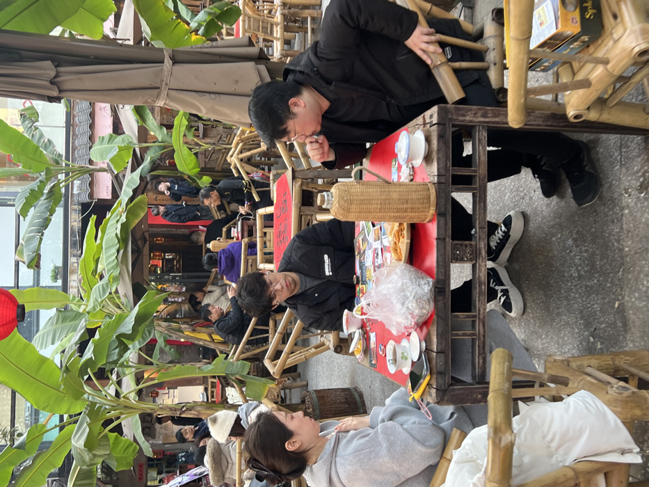 A scene in Yuzhong District, China, depicts an outdoor dining or tea house area bustling with activity under natural daylight. In the foreground, three individuals are seated around a rustic, red-topped wooden table with a dark wooden base, on a concrete surface.
On the left, a woman with dark hair secured by a clip wears a light gray hooded sweatshirt. She is looking downwards, potentially at a yellow-cased mobile phone visible on the table, with her left hand near her chest. Next to her, a man with glasses and short dark hair, wearing a dark puffer jacket and white wireless earbuds, looks downwards with a neutral expression. On the right, another man, also wearing glasses and a dark puffer jacket, looks towards the middle man, with his right hand positioned near his mouth.
The table is laden with multiple items, including small white teacups/bowls, a clear plastic bag containing numerous small, silver-colored objects (possibly dried fish or snacks), a wicker-wrapped thermal flask, and a red plate holding golden-brown snacks. A red rectangular object with Chinese characters is also visible on the table. All three individuals are seated on bamboo chairs.
The wider environment features numerous large-leafed tropical plants resembling banana trees, providing greenery and partial shade. Large beige outdoor umbrellas are deployed over some seating areas. In the background, many other patrons are seated at similar bamboo tables, engaged in social interaction or dining. Traditional Chinese architectural elements, such as red lanterns and dark wood storefronts, line the perimeter, indicating a commercial area or a traditional pedestrian street. A single red lantern hangs in the upper left of the frame.