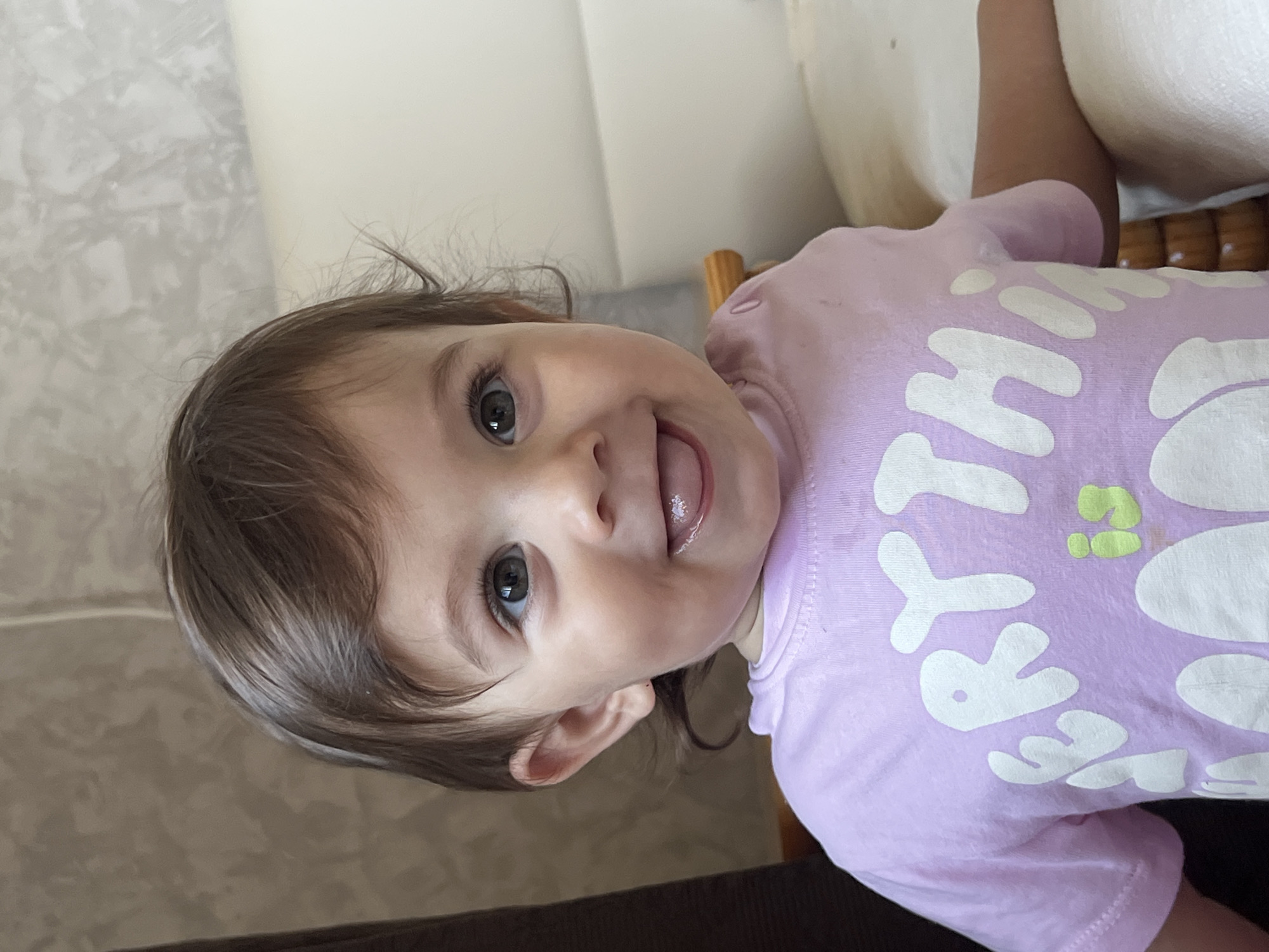 An overhead medium shot captures a baby girl looking up and to her right, with her tongue sticking out. She is wearing a light purple t-shirt with white lettering. The background consists of a textured wall and a light-colored couch or bed. The lighting is bright, and the focus is sharp on the baby's face. No specific location in Elena, Bulgaria can be determined from the image.