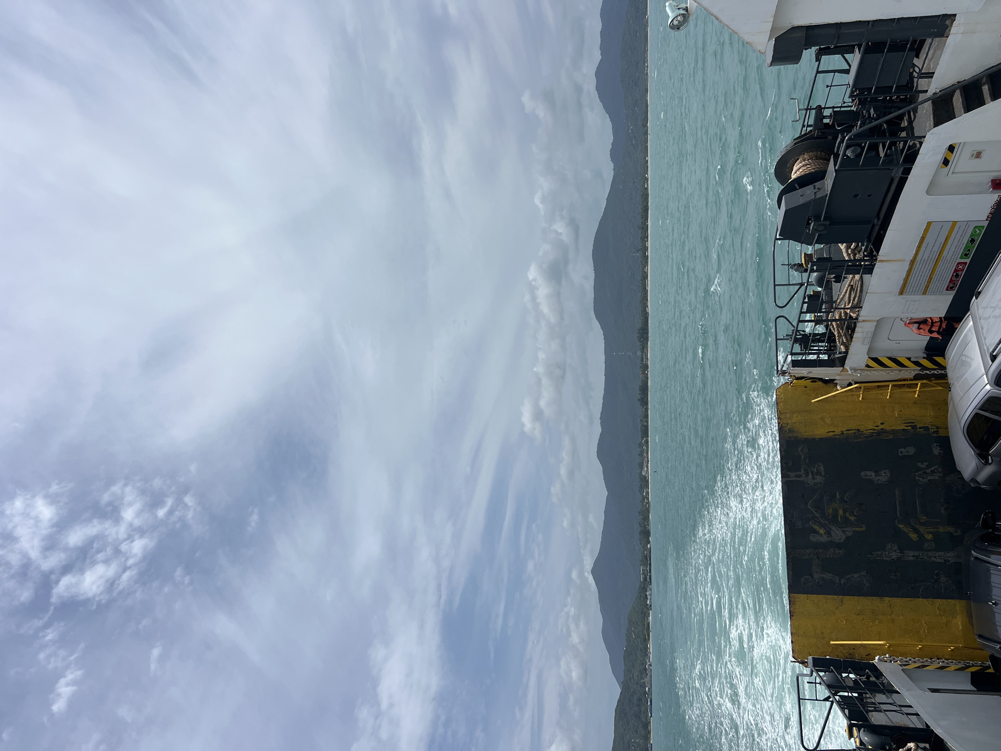 This image depicts the rear deck of a ferry crossing the turquoise waters off the coast of Ko Pha-ngan District, Thailand. The ferry is actively moving, indicated by the frothy wake trailing behind. Several vehicles, including a dark SUV and a silver sedan, are visible loaded onto the deck. Prominent on the deck is a large, weathered yellow and black ramp. The surrounding environment shows a coastline with lush green mountains and a narrow strip of sandy beach. The sky is overcast with wispy white clouds. The water is choppy with visible waves.