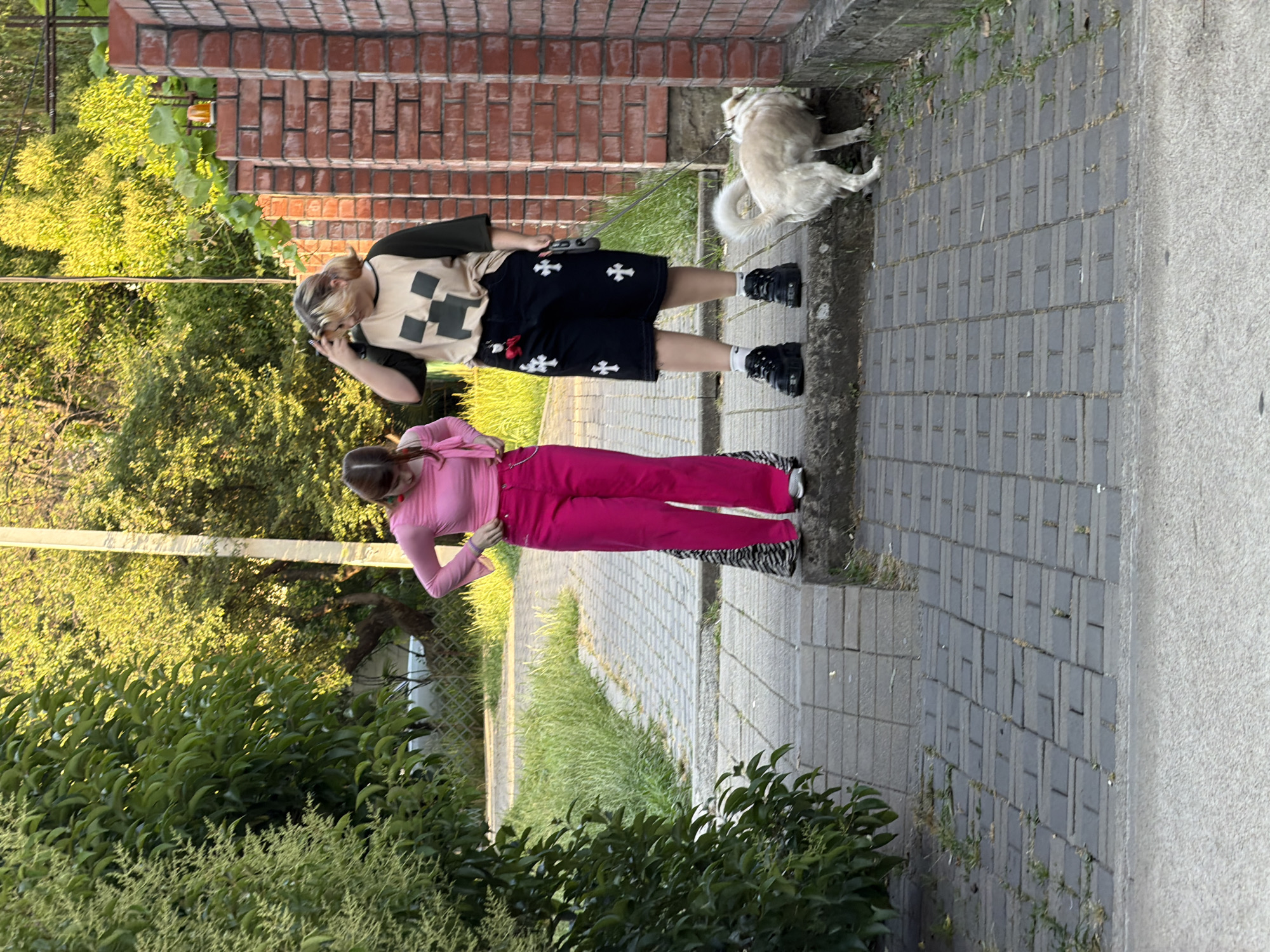 This image depicts two young women and a dog on a paved walkway lined with brick walls and lush green foliage. The woman on the left is wearing a bright pink long-sleeved shirt and hot pink wide-leg pants. She has her hair pulled back in a ponytail and is holding a pink purse. The woman on the right is wearing a beige t-shirt with a black Minecraft Creeper design and a black skirt adorned with white cross symbols. She has her hair down and is touching her head. She is holding a leash attached to a light-colored, fluffy dog. The dog is sniffing at the base of the brick wall. The environment appears to be outdoors, possibly a residential area or park, with trees and a lamppost visible in the background. The walkway is made of interlocking gray pavers. There is no specific indication of the city of Sochi, Russia within the image.