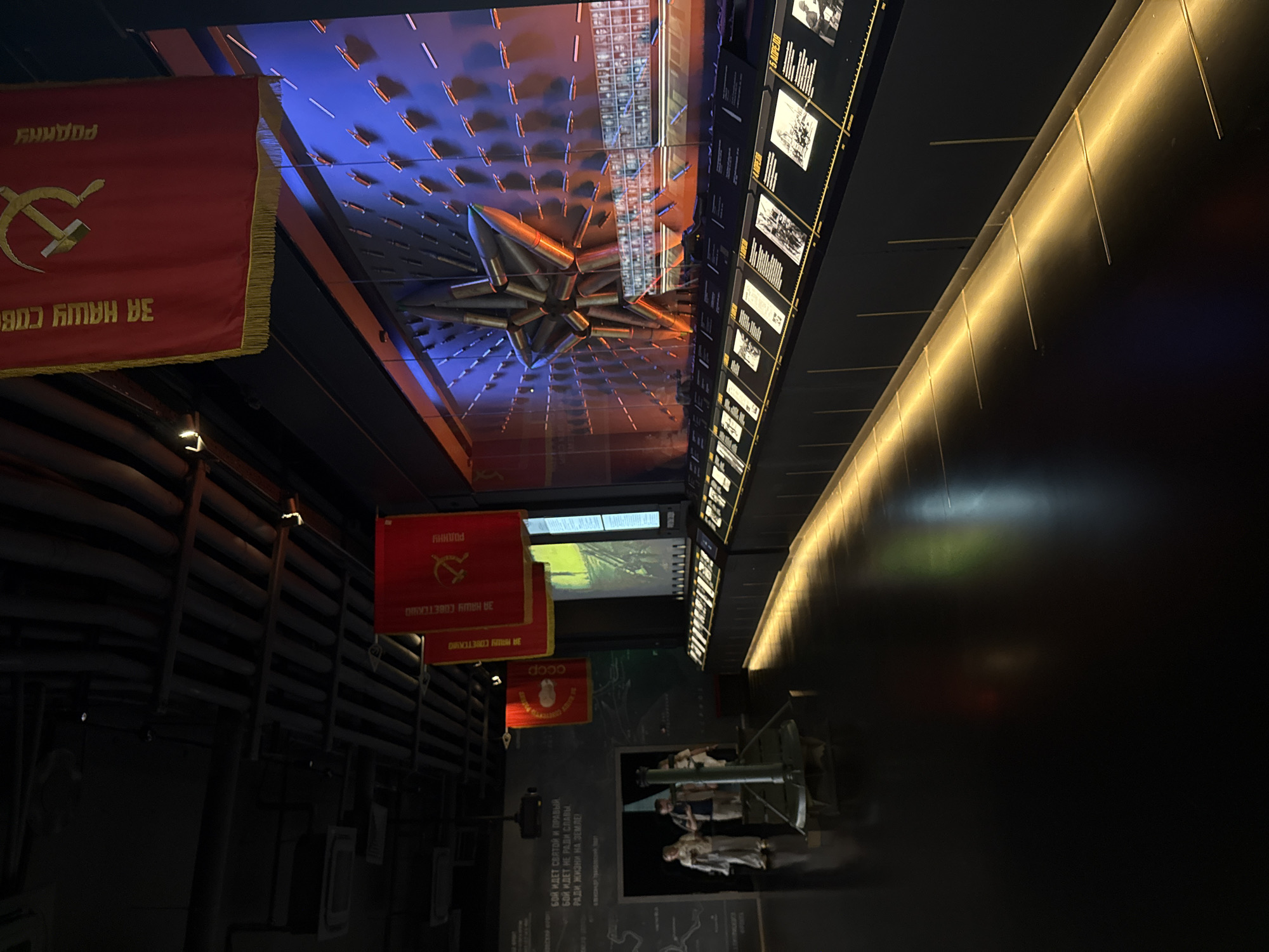 This image depicts the interior of a museum exhibit, likely a war memorial or historical exhibition, in Zelenaya Roshcha, Russia. The scene is dimly lit with strategic spotlights illuminating various displays. In the foreground, the polished dark floor reflects the ambient light.
Several people are visible in the middle ground, standing near an entrance. One individual is looking at a display on the wall. Two other individuals are walking away from the camera. In front of them, a large, dark-colored object, possibly a cannon or mortar, is positioned on the floor.
The dominant feature of the background is a large, illuminated wall display. This display is dramatically lit with blue and orange hues, highlighting a prominent, large star made of what appear to be artillery shells or similar metallic objects. The star is surrounded by rows of smaller shells or projectiles mounted on the wall, creating a textured and imposing visual.
Above the main display, red banners with yellow trim are hung from the ceiling. These banners feature Cyrillic lettering and Soviet-era symbols, such as a hammer and sickle. The ceiling itself is dark and appears to be an industrial design with exposed pipes and ventilation systems.
Along the bottom of the large wall display, a row of illuminated exhibit panels is visible, each containing text and images. These panels are arranged linearly, suggesting a chronological or thematic presentation of historical information. The overall atmosphere is solemn and commemorative, befitting a museum dedicated to historical events.