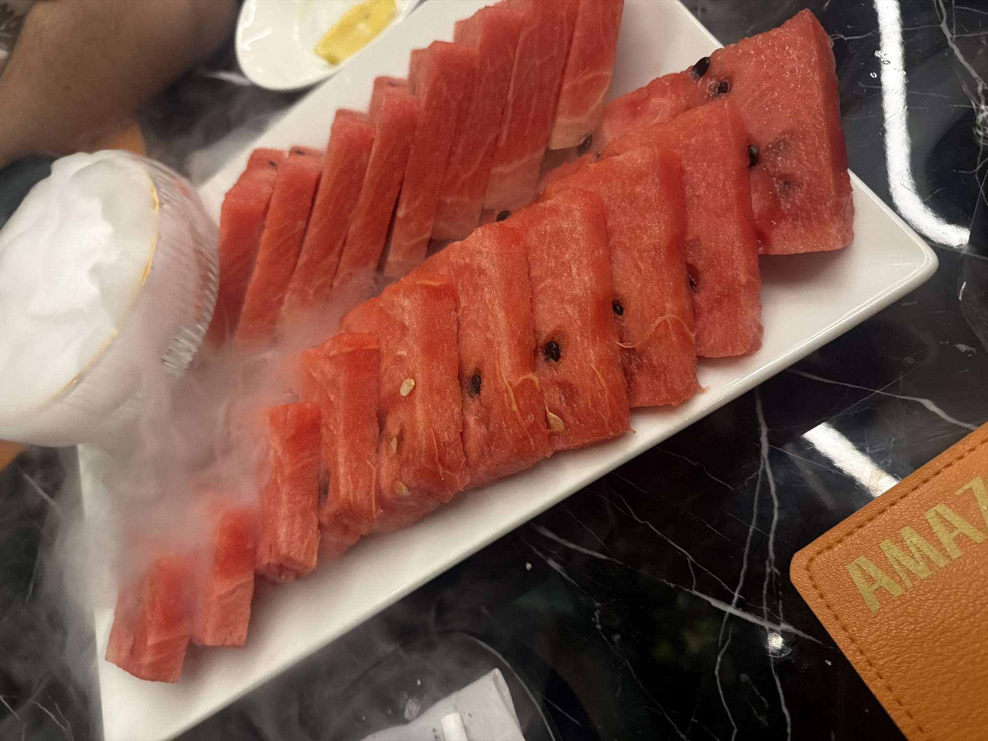 A platter of sliced watermelon is presented on a white, rectangular serving dish. The watermelon is arranged in two rows and appears to be emitting a white vapor. A small dish of sliced fruit, potentially banana or lemon, is also visible. To the right of the platter, a brown object with the word "AMAZ" printed on it is visible. The surface underneath the platter appears to be a black marble tabletop. The origin is likely the city of Yerevan, Armenia.