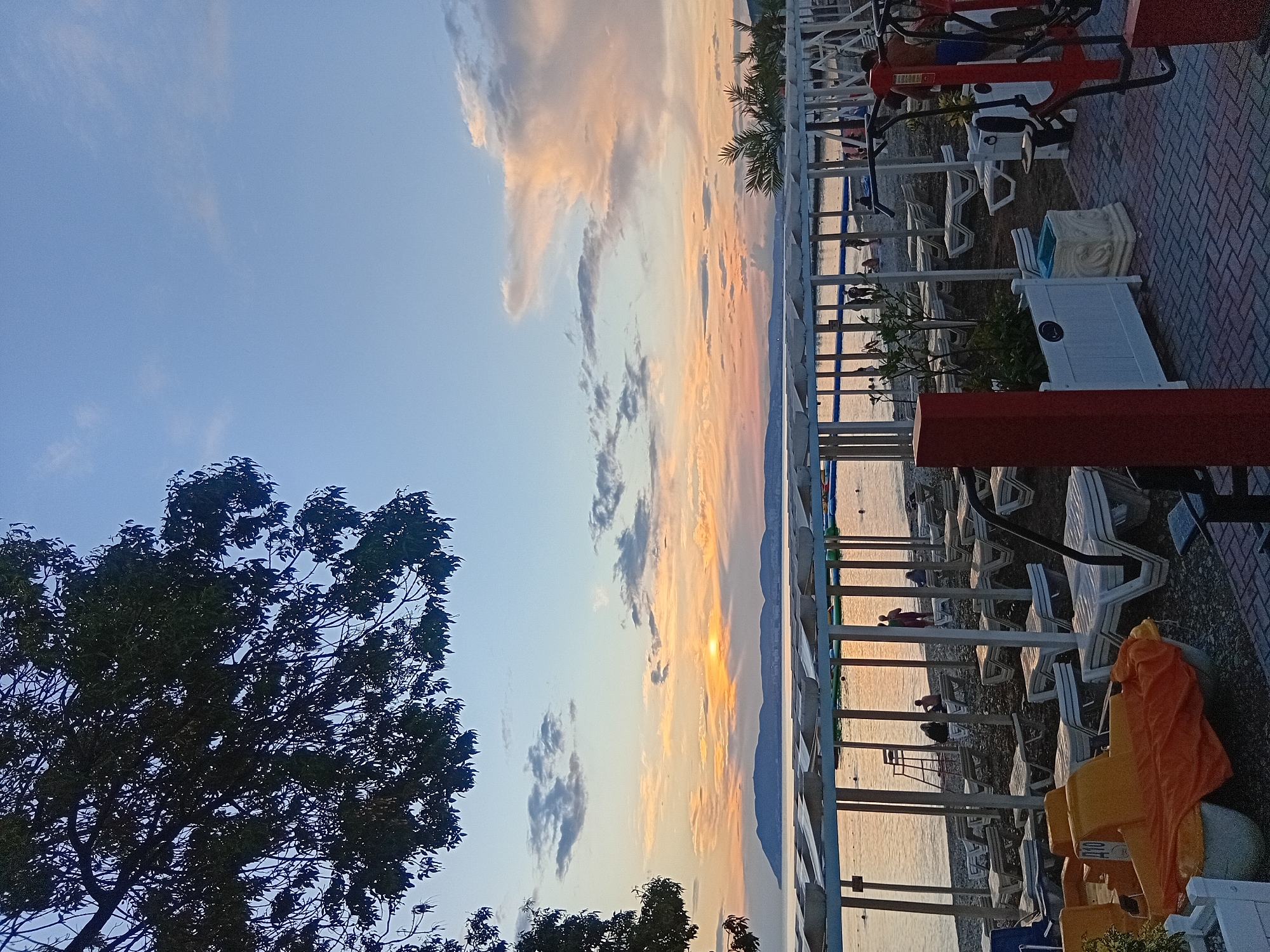 The image depicts a beach scene at sunset in Kirillovka, Russia. In the foreground, there are several yellow pedal boats, some covered with orange tarps, on a pebble beach. Further back, rows of empty white lounge chairs are arranged under a white canopy. The sea is calm and reflects the warm colors of the sunset. In the middle ground, people are visible walking along the shoreline and in the water. A lifeguard tower stands near the water's edge. In the background, a distant mountain range is silhouetted against the colorful sky, which features hues of orange, yellow, and pink from the setting sun. Trees with dark foliage frame the upper left portion of the image. A portion of outdoor exercise equipment is visible in the lower right corner.