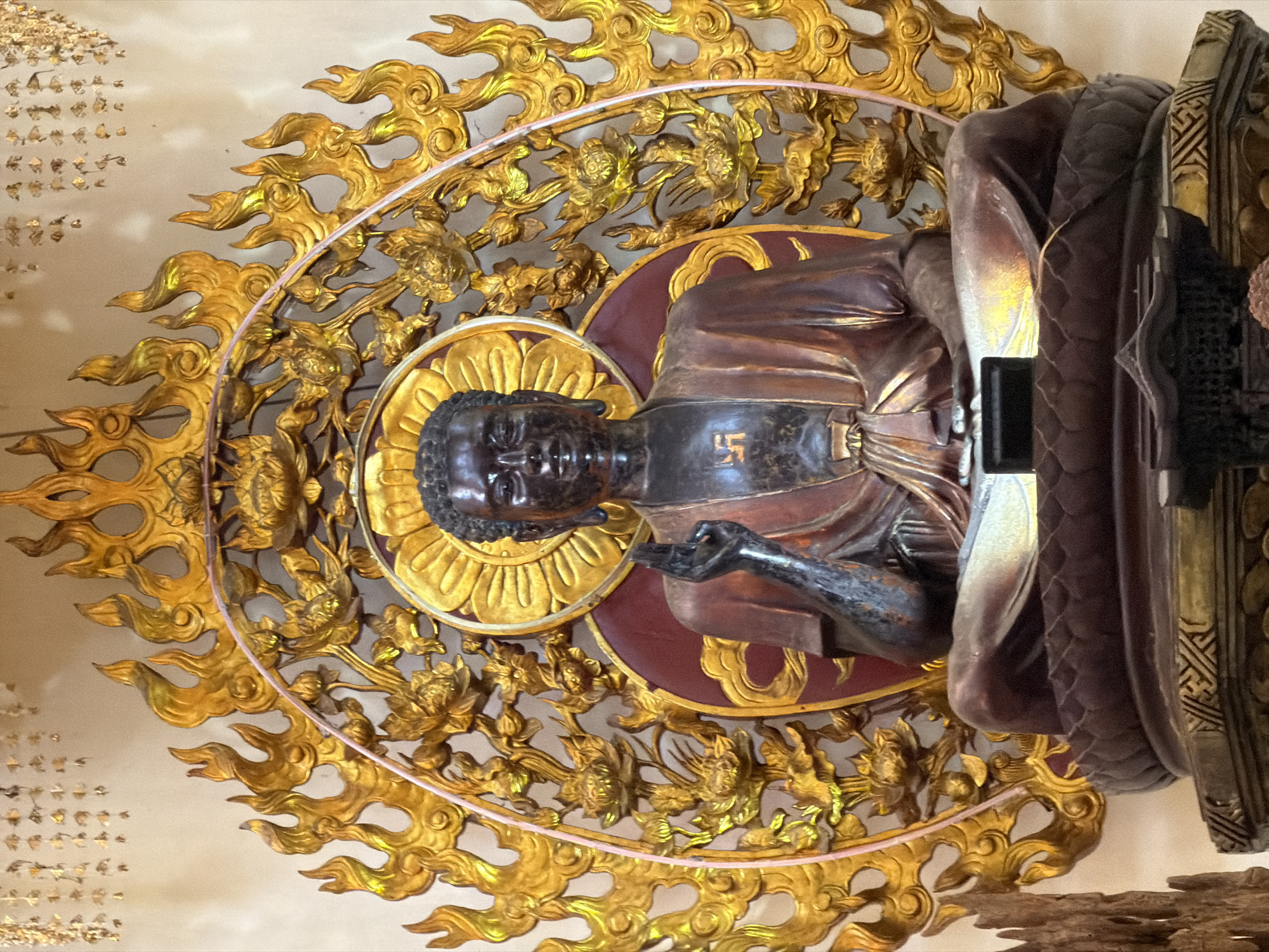 A full-shot, indoor, rotated view displays a Buddhist statue in Nha Trang Ward, Vietnam. The statue is bronze, decorated with gold and is positioned in front of a wall with numerous gold details. The Buddha is seated, with his right hand outstretched, and has a golden halo. On the Buddha's chest is a Swastika. He is surrounded by an oval-shaped ring of golden, flame-like designs. The statue is elevated, suggesting it is on display. Light is reflecting off the golden components, giving the scene a warm, vibrant tone.