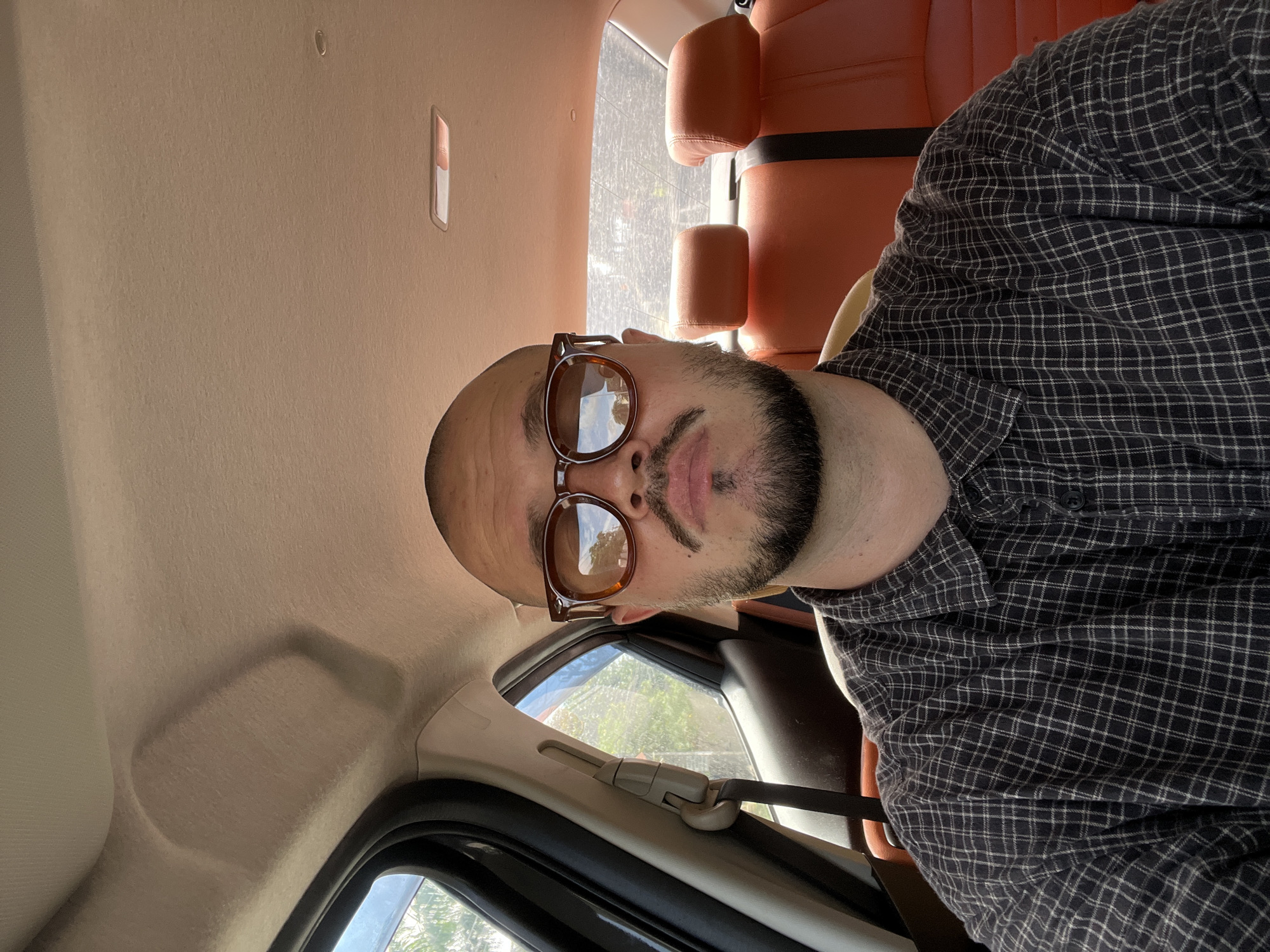 The image is a close-up, frontal, outdoor selfie of a bald man with a beard and mustache wearing brown-tinted sunglasses and a dark plaid shirt. He is seated in the back of a car with orange leather seats. The interior of the car is visible, including the headliner and a window on the left side, through which greenery and a bright sky are seen. The man is the sole subject of the image. The overall lighting suggests it is daytime.