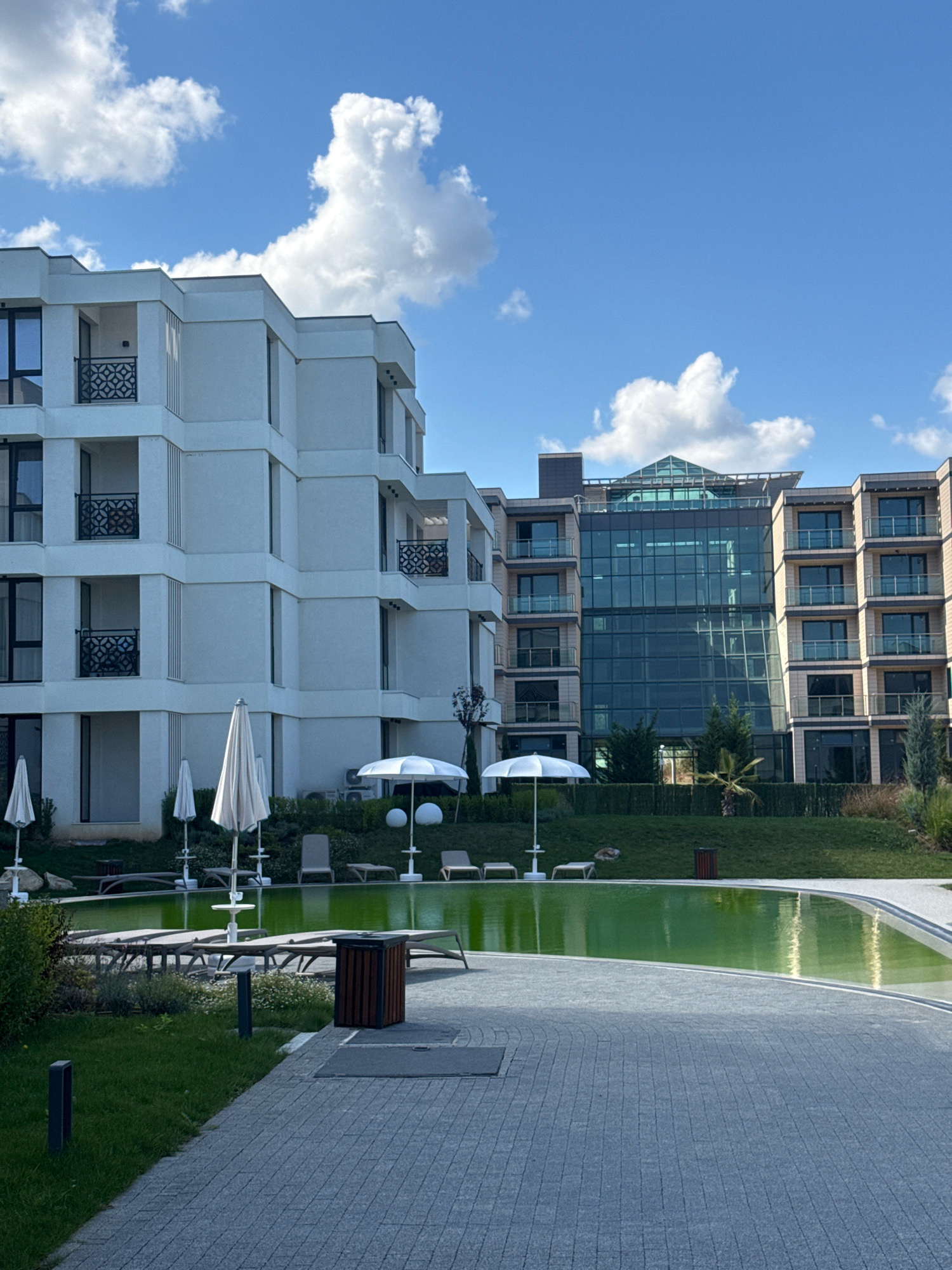 The image shows a modern resort complex with white residential buildings and a central glass-fronted commercial building. In the foreground, a paved walkway leads to a kidney-shaped swimming pool filled with vibrant green water. Lounge chairs and white umbrellas are scattered around the pool area, on a neatly manicured lawn. The buildings feature clean lines and balconies with black metal railings. The sky is a clear blue with scattered white clouds. The scene suggests a well-maintained leisure facility, likely a hotel or a residential complex, located in or near Sozopol, Bulgaria. There are no people visible in the image.