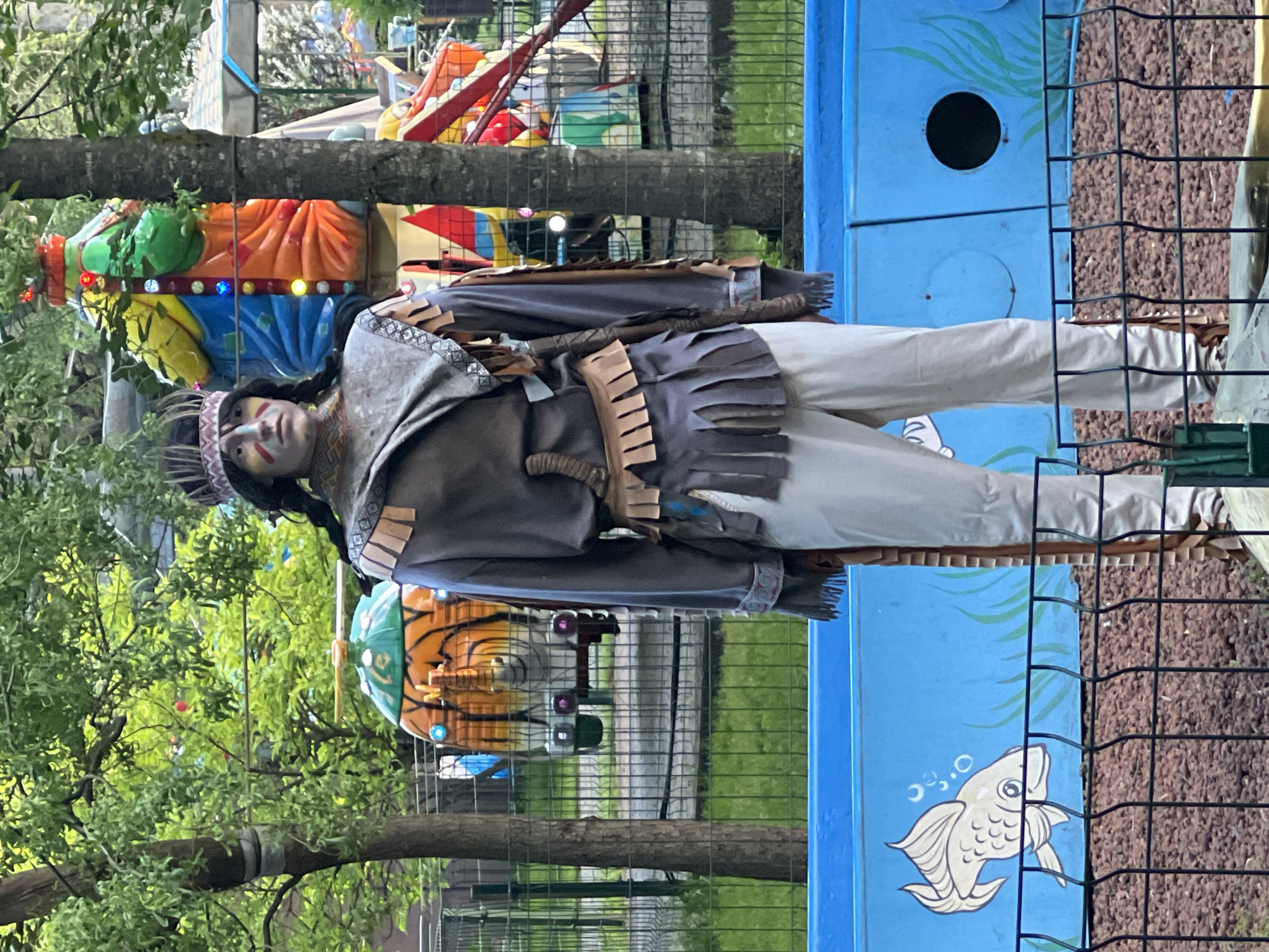 A life-size Native American mannequin stands outdoors in a park, likely in Krasnodar, Russia. The mannequin is dressed in traditional Native American attire, including a fringed brown jacket, light-colored pants, and a feathered headband. The mannequin has painted face markings. It is positioned next to a blue structure with aquatic-themed decorations. A black metal fence is partially visible in the foreground. Trees and amusement park rides are in the background.