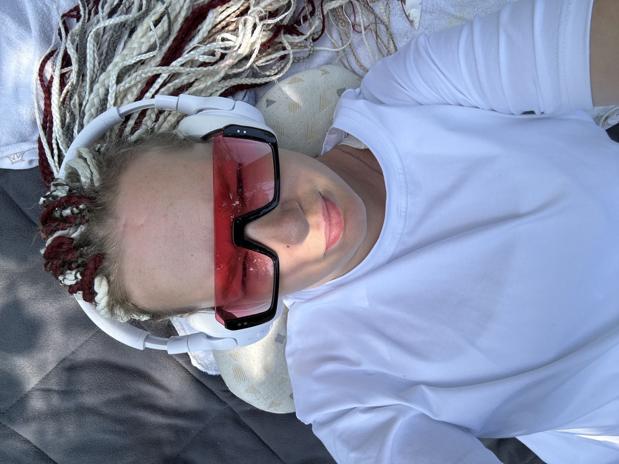 A person lies on their back, facing the camera, with their head on a light-colored patterned pillow. They are wearing white headphones with red-tinted sunglasses. Their hair is styled in numerous braids, some appearing to be reddish-brown and white. The person is also wearing a plain white t-shirt. The background consists of a textured dark gray surface, possibly a blanket or mat, and a white towel. The lighting suggests an outdoor environment with sunlight casting shadows. There are no visible Moscow landmarks.