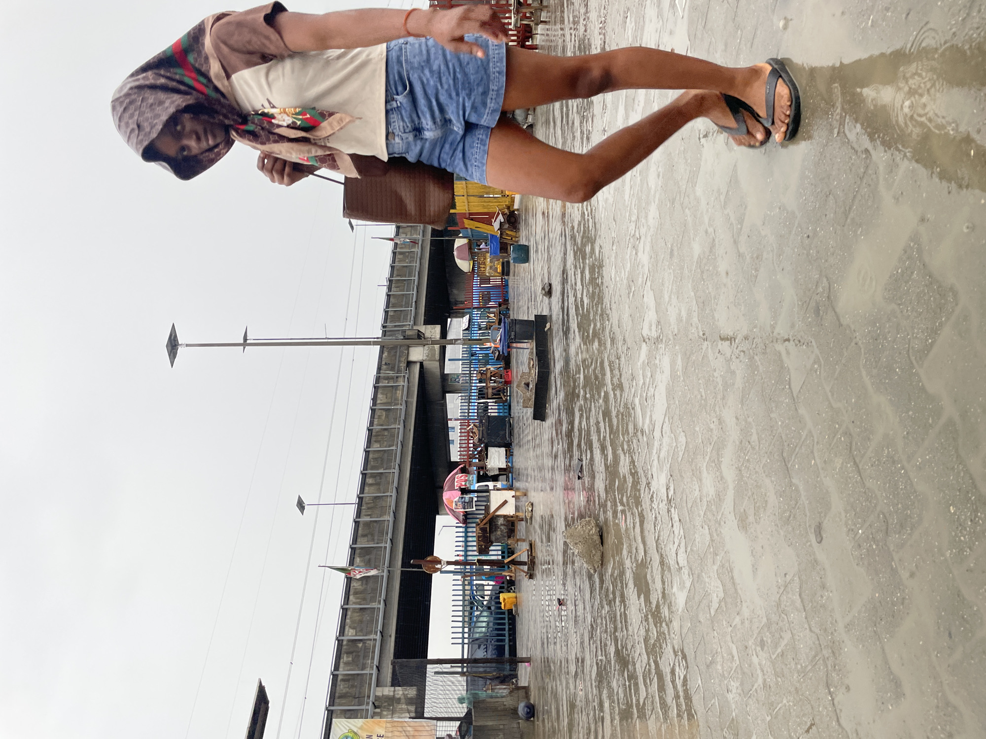 A young individual stands on a wet, paved surface in Lagos, Nigeria. They are wearing denim shorts, a light-colored shirt, a patterned headscarf, and brown flip-flops. The individual is positioned in front of a bustling market area under a concrete overpass. Market stalls are visible, with various objects and people present. The sky is overcast. The paved surface appears to be wet from rain, with reflections and small puddles visible.