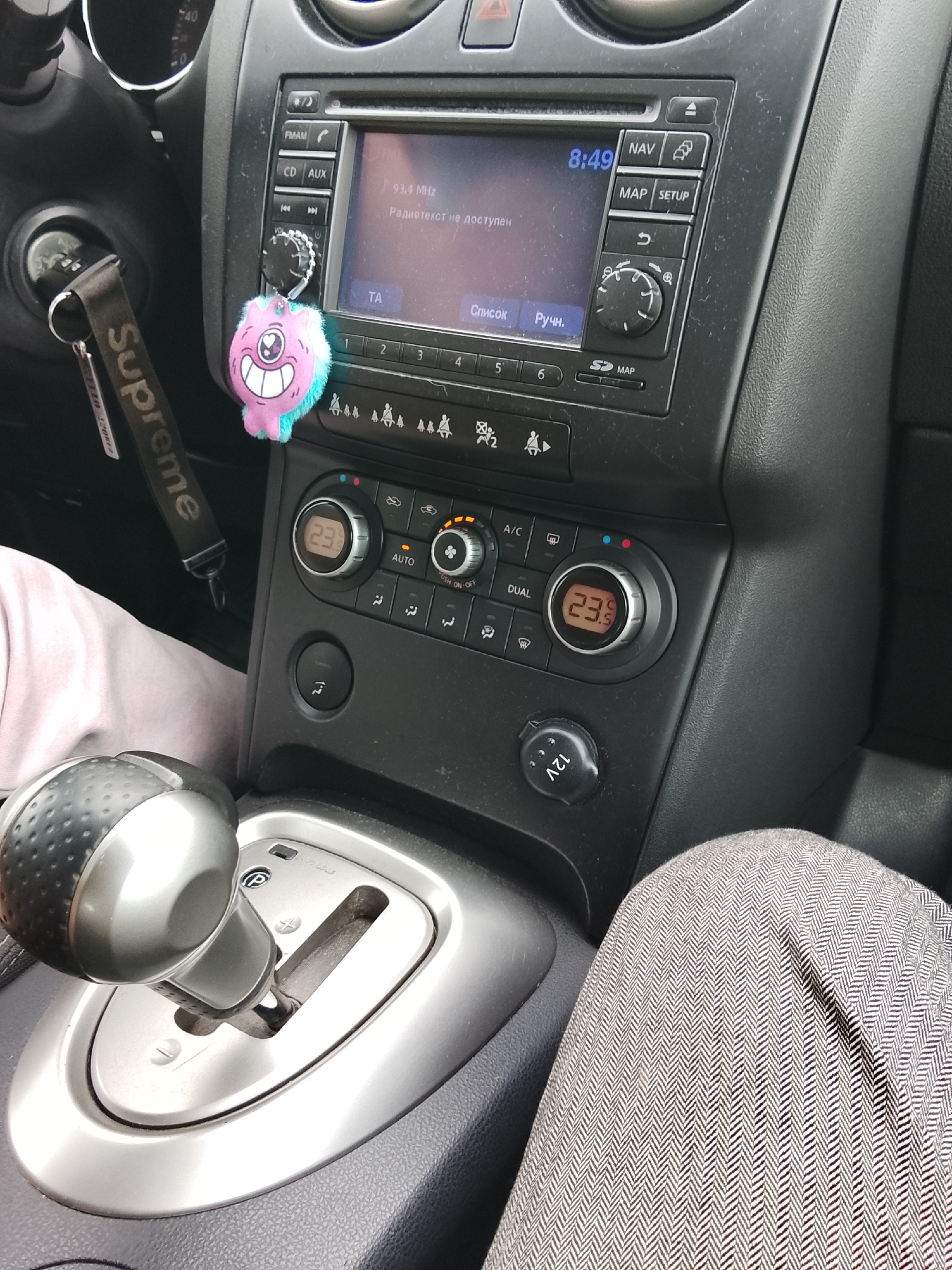 The image shows the interior of a car, likely a Nissan Rogue based on the dashboard features. The gear shift is in the foreground. A leg is visible wearing pants with a herringbone pattern. A "Supreme" keychain hangs from the ignition. The car's radio displays "93.4 MHz" and a message in Russian, "Радиотекст не доступен" (Radiotext not available). The clock reads 8:49. The climate control system is set to "AUTO" and a temperature of 23.9. The location is Высокиничи, Russia.
