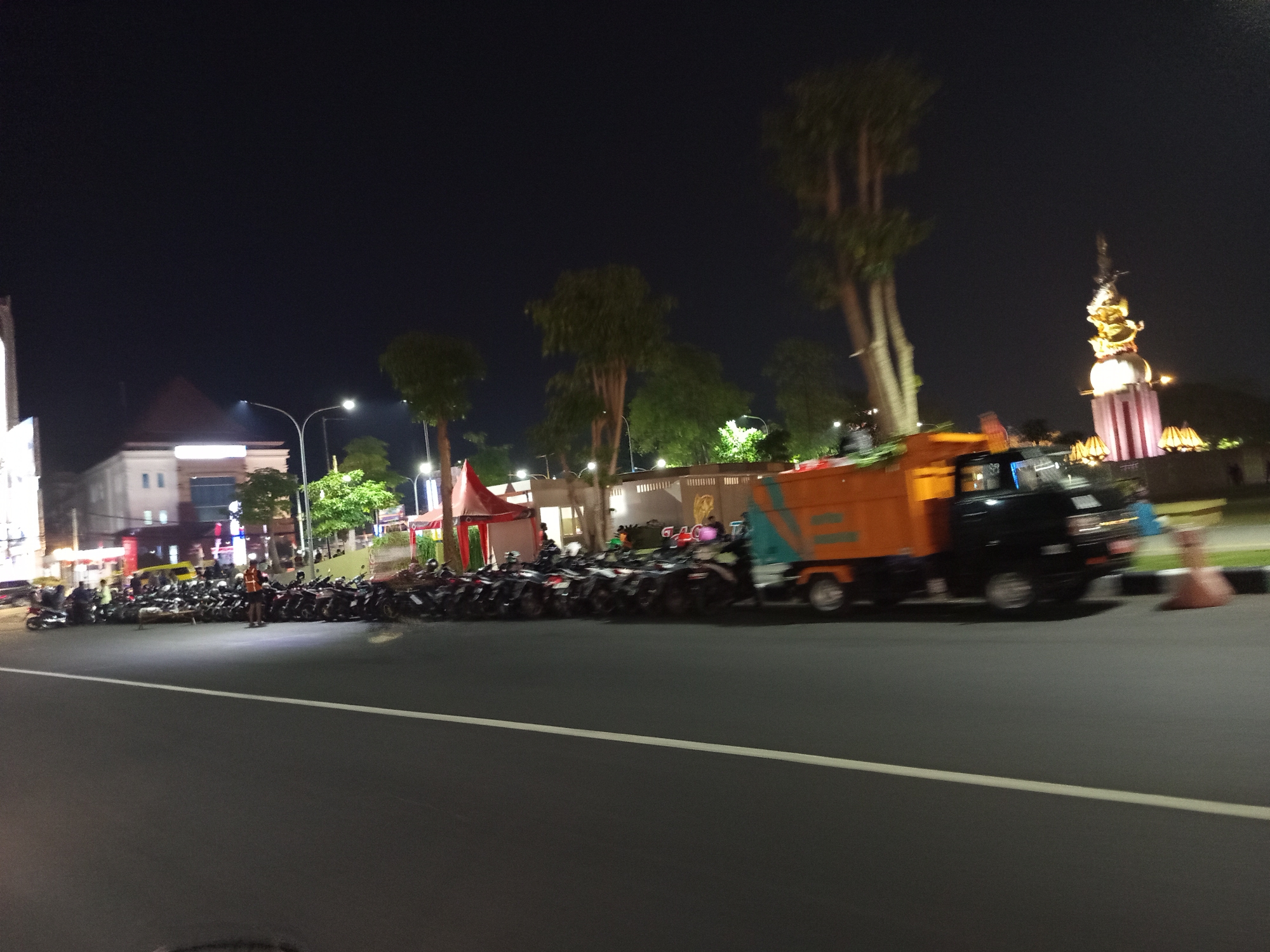 This is a night-time photograph of a street scene in Sidoarjo, Indonesia. On the right side of the image, a dark colored truck with an orange bin is driving on the road. In front of the truck, there are traffic cones. To the left of the truck, a large number of motorcycles are parked along the side of the road. Behind the motorcycles, there are several trees and buildings illuminated by lights. One building appears to be a mosque with a tall, golden dome. The sky is dark and clear. There are streetlights visible throughout the scene. A white line marks the edge of the road in the foreground.