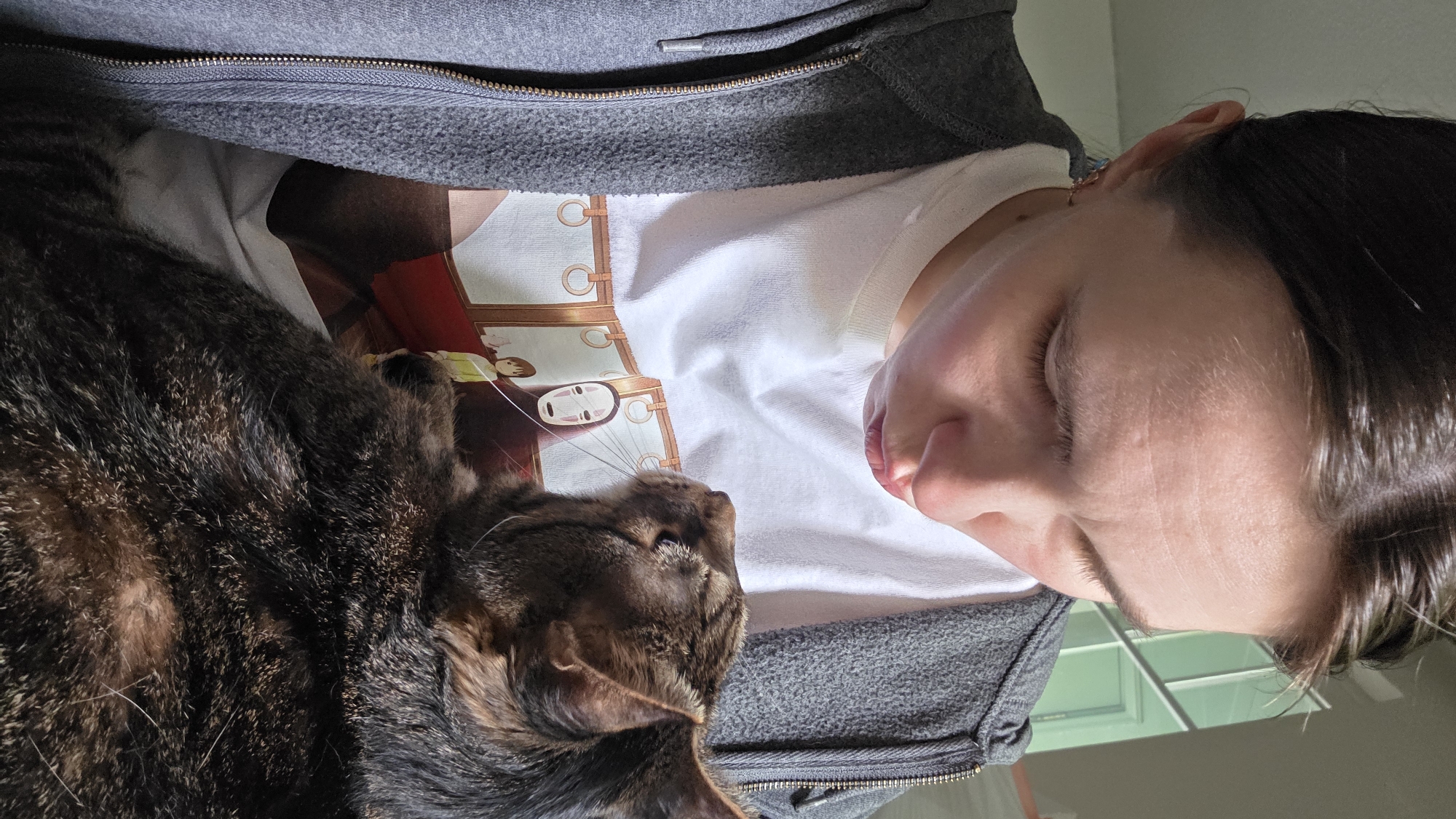 A person, identified as female, is shown from the chest up, looking down at a cat held in her arms. She has short, dark brown hair and is wearing a grey zip-up hoodie over a white t-shirt with a graphic print. The t-shirt depicts a scene from the anime "Spirited Away," featuring the character No-Face and Chihiro on a train. The cat is a tabby with dark brown and black fur. The person's face is angled downwards, with her lips pursed. The cat's head is tilted upwards, looking towards the person. The background is out of focus, but appears to be an indoor setting with a window visible. The image is a close-up shot, emphasizing the interaction between the person and the cat. The location is not specified in the image.