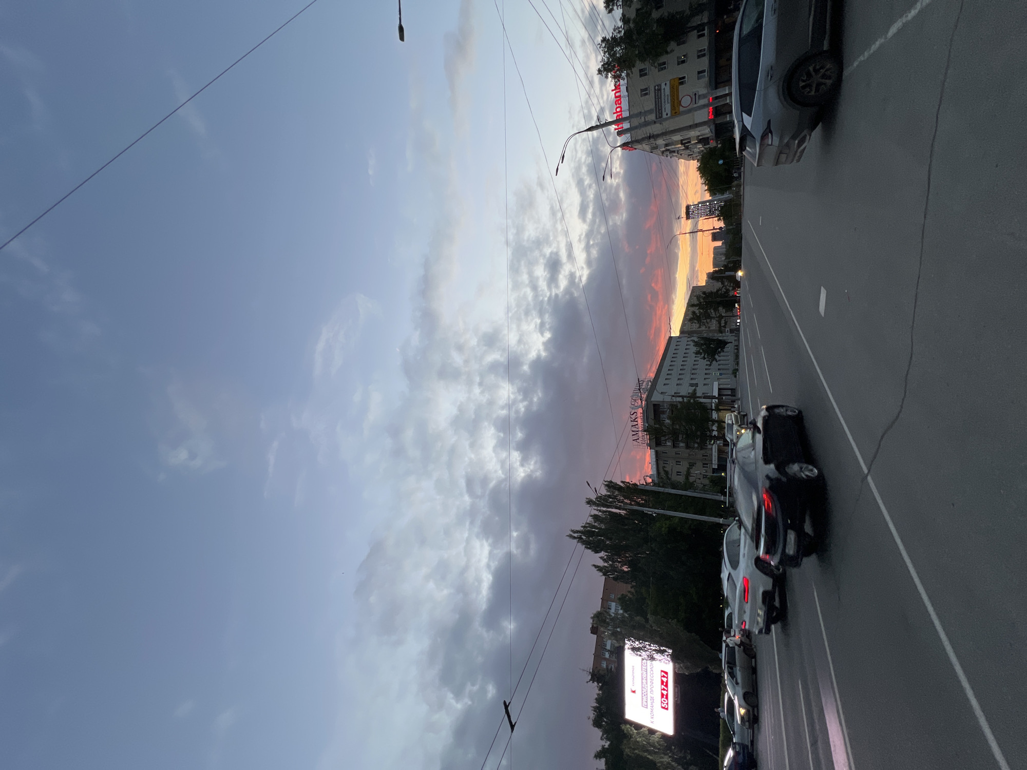 The image depicts a street scene at dusk in Izhevsk, Russia. The sky is filled with dramatic clouds tinged with the colors of sunset, ranging from pale blue to hues of pink and orange. Overhead, power lines crisscross the frame. A multi-lane road is visible in the foreground, with several cars driving or stopped.
On the left side of the road, a large digital billboard displays text in Russian, including "ПРИСОЕДИНЯЙТЕСЬ К КОМАНДЕ ПРОФЕССИОНАЛОВ" which translates to "JOIN THE TEAM OF PROFESSIONALS," and a phone number. Adjacent to the billboard are trees and buildings.
On the right side of the road, several multi-story buildings line the street. One building has a red sign with Russian text, appearing to be a business or advertisement. Another building has signage with the word "AMAKS" and possibly "CENTR." A street lamp is visible atop one of the buildings. The architecture suggests a typical urban environment. The overall impression is of a city winding down for the evening, with the natural beauty of the sunset providing a striking backdrop to the urban landscape.