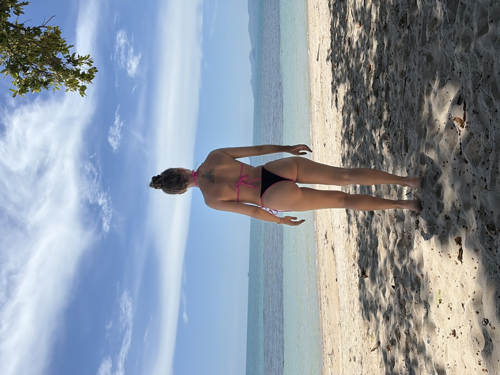 A young woman stands on a sandy beach, facing away from the camera, looking out at the ocean. She is wearing a black bikini with pink trim. Her hair is pulled up in a bun. In the background, the ocean stretches to the horizon, with a clear blue sky above. The beach is scattered with fallen leaves and branches, and shadows from unseen trees fall across the sand. The location is identified as Baan Tai, Thailand.