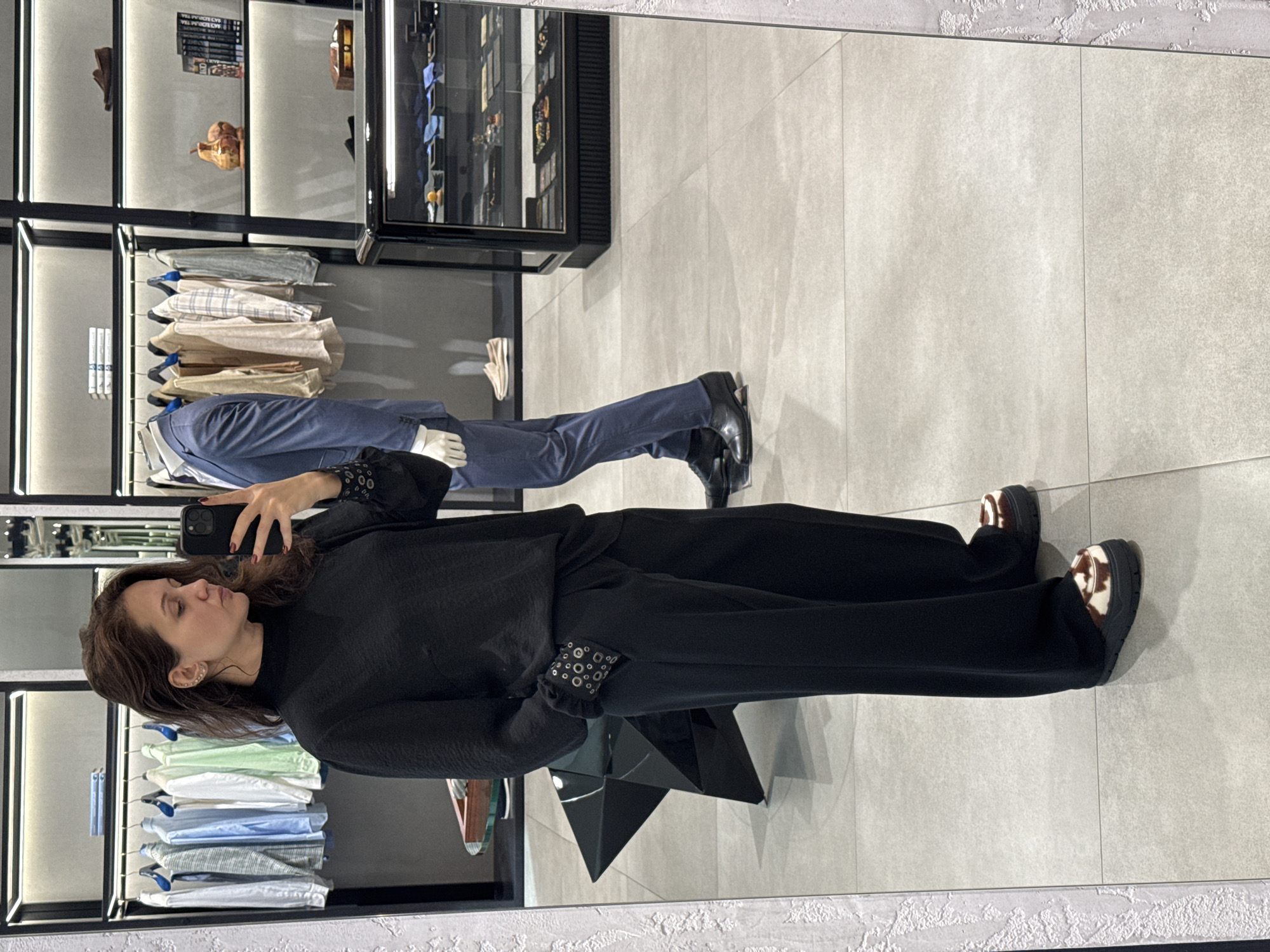 A woman takes a picture of herself in a large mirror inside a clothing store. The reflection shows a mannequin dressed in a blue suit, shirts on hangers, and merchandise in glass cases. The floor is light gray tile. The scene appears to be indoors, possibly in a high-end retail location. It is not possible to definitively state this is Dubai, United Arab Emirates from visual elements alone.