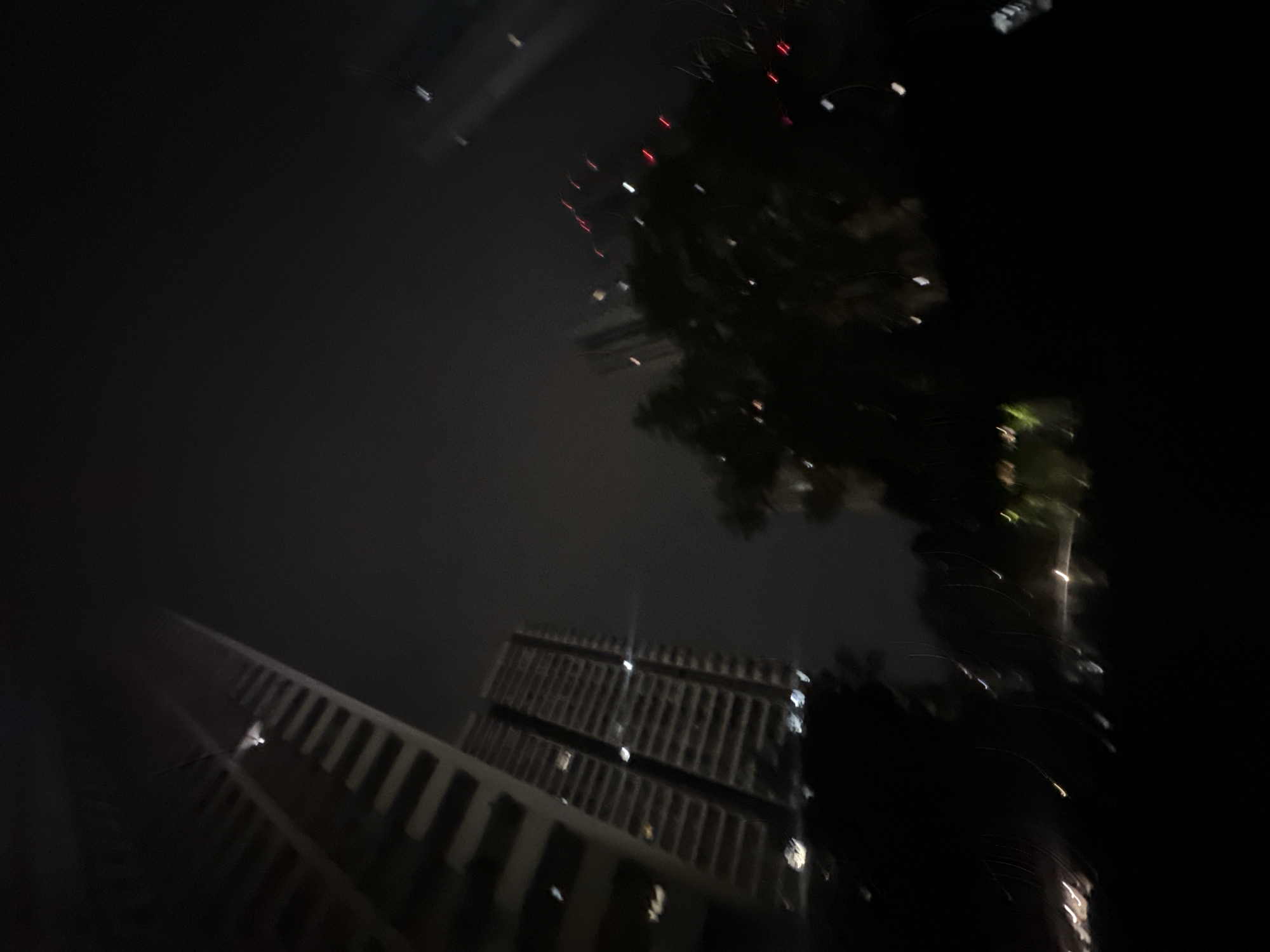 This is a low-light, motion-blurred photograph depicting an urban environment at night in the Chongqing High-tech Zone, China. The scene is dominated by multiple tall buildings and dark vegetation under a dark, possibly hazy, sky.
On the far left, a large high-rise building is partially visible, showing horizontal architectural elements and a prominent, blurred light source near its upper section. Towards the center-left, a second multi-story building is discernible with several illuminated windows appearing as bright, elongated blurs. Dark, indistinct masses representing tree canopies occupy the foreground and mid-ground, obscuring the lower portions of the structures.
Throughout the image, numerous artificial light sources are present as streaks and scattered blurs of white, yellow, and red, consistent with distant building lights, streetlights, or vehicle movement, intensified by the motion blur. The overall impression is one of an active urban area captured with a long exposure or significant camera movement. No human subjects, distinct actions, or specific interactions are identifiable.