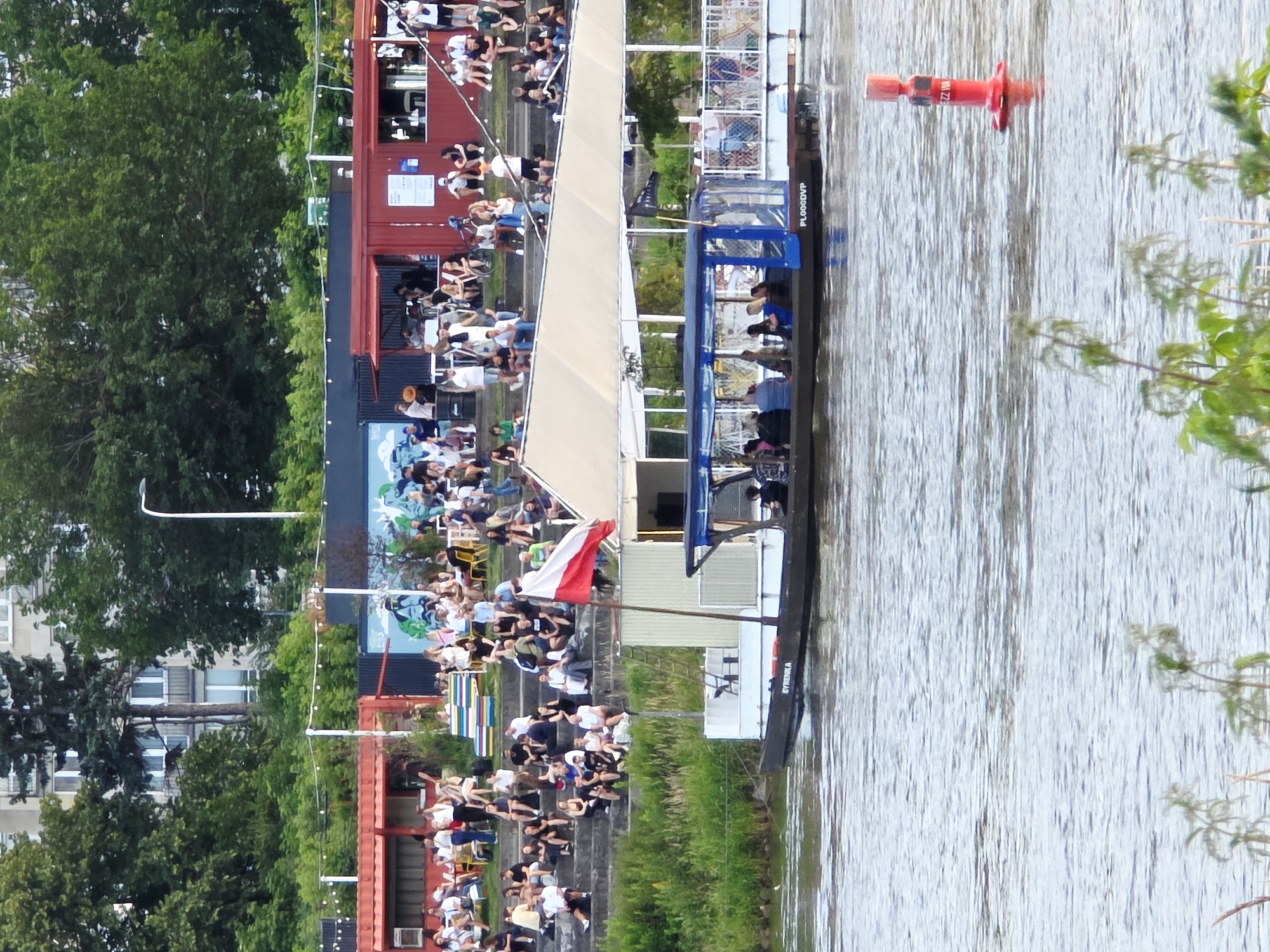 The image depicts a vibrant waterfront scene in Warsaw, Poland. In the foreground, a boat with a covered seating area is navigating the river, carrying passengers. The boat has the name "SYRENKA" visible on its hull, along with a registration number "PL000DVP". A red buoy is also visible in the water.
The background reveals a lively embankment lined with what appear to be shipping containers converted into shops or cafes. A diverse crowd of people is seated on concrete steps leading down to the river, enjoying the atmosphere. Some are socializing, others are seated on the steps, and a few are standing. A Polish flag is prominently displayed on a flagpole on the boat. Lush greenery and trees frame the scene, with buildings visible in the distance behind the trees, characteristic of an urban environment. The overall impression is one of a popular recreational area where people gather to relax and enjoy the waterfront.