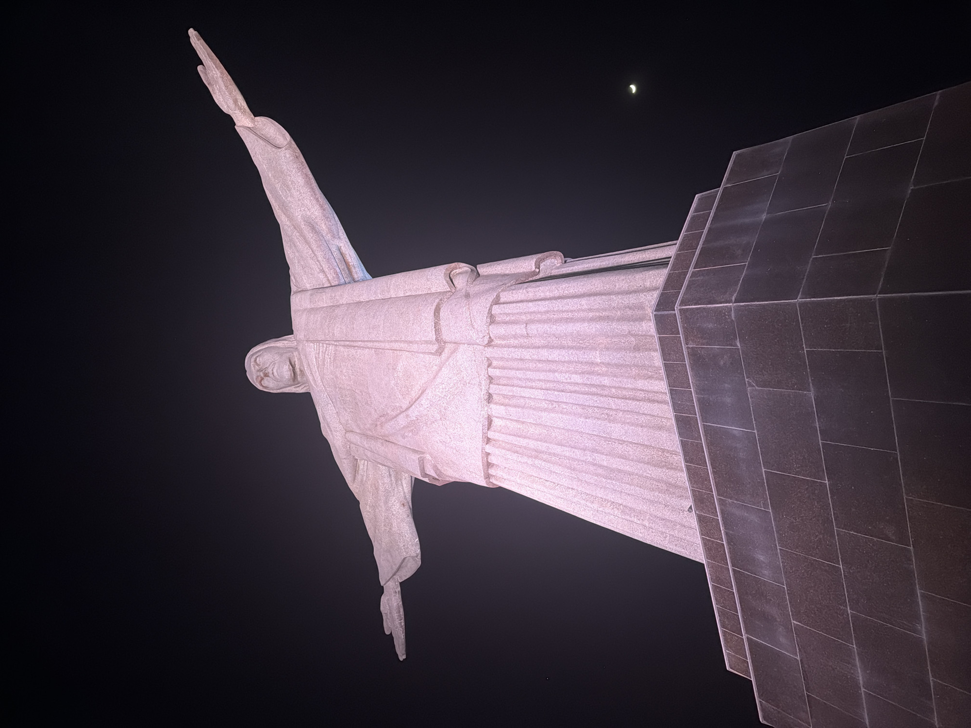 The image shows a low-angle, nighttime shot of the Christ the Redeemer statue in Rio de Janeiro, Brazil. The statue, made of stone, is illuminated and stands against a dark night sky. The iconic figure's arms are outstretched, and its robe is depicted with folds and pleats. The statue is situated on a large, multi-tiered stone base. A crescent moon is visible in the upper right quadrant of the sky, along with a faint, distant light that could be a star or a plane. The overall impression is one of grandeur and solemnity.