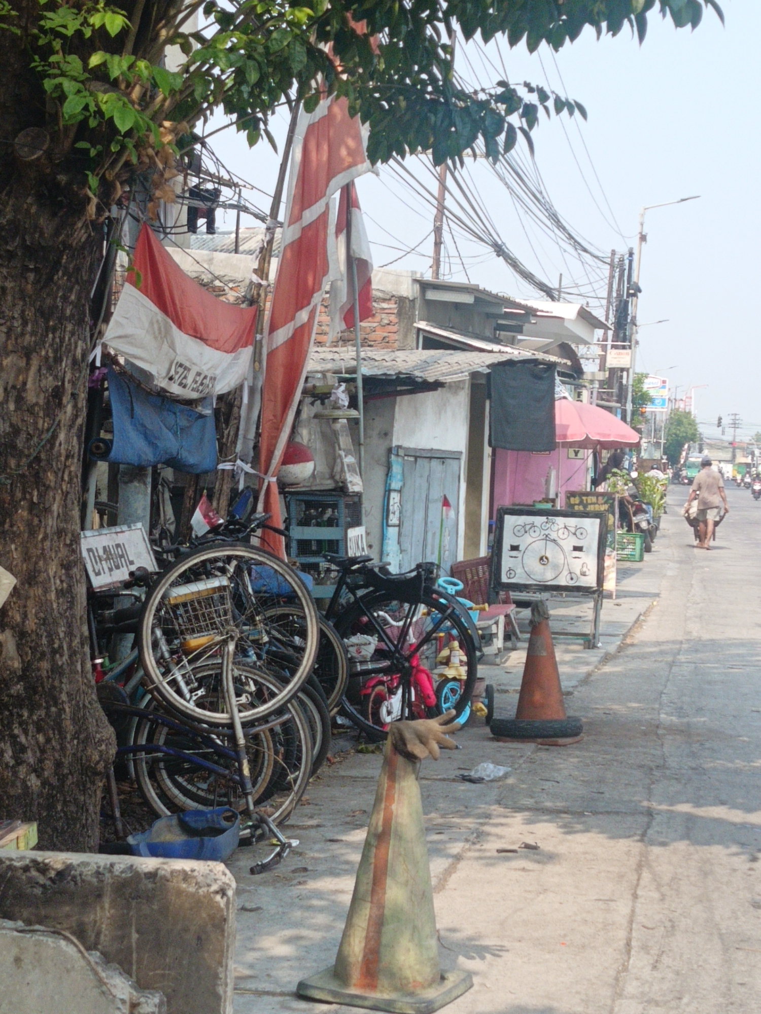 The image depicts a street scene in Gedangan, Indonesia, with a focus on a collection of bicycles parked near a building. A large tree with lush green leaves stands on the left, partially obscuring a building with a corrugated roof and brickwork. Indonesian flags, red and white, are displayed prominently, one draped over a pole and another partially visible. Several bicycles are stacked and leaning against each other, some with visible baskets and frames. A sign with "DI-JUAL" (For Sale) is attached to a bicycle. A blue plastic object lies on the ground near the bicycles. In the background, power lines crisscross overhead, and more buildings and a street with a few people and vehicles are visible. One person, dressed in a light-colored shirt and shorts, is walking away from the camera down the street, pushing a cart. The scene suggests a commercial area or a residential street with a business involving bicycles.
