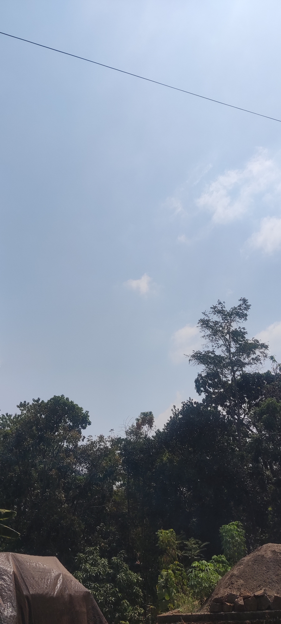A clear blue sky with sparse white clouds stretches across the top of the frame. A thin, dark wire runs diagonally from the upper left corner to the mid-right of the image. Below the sky, a dense canopy of dark green trees forms a natural horizon. Among the trees, a tall, slender tree with lighter green foliage stands out on the right side. In the lower left corner, a tarp-covered object is visible, suggesting some form of outdoor storage or construction. The bottom right of the image shows a pile of dark brown soil or dirt, possibly part of a construction site, with some rough-textured bricks at its base. The scene appears to be an outdoor location, possibly in Panongan Lor, Indonesia, given the natural foliage and the appearance of unfinished construction elements. There are no people or animals visible in the image.