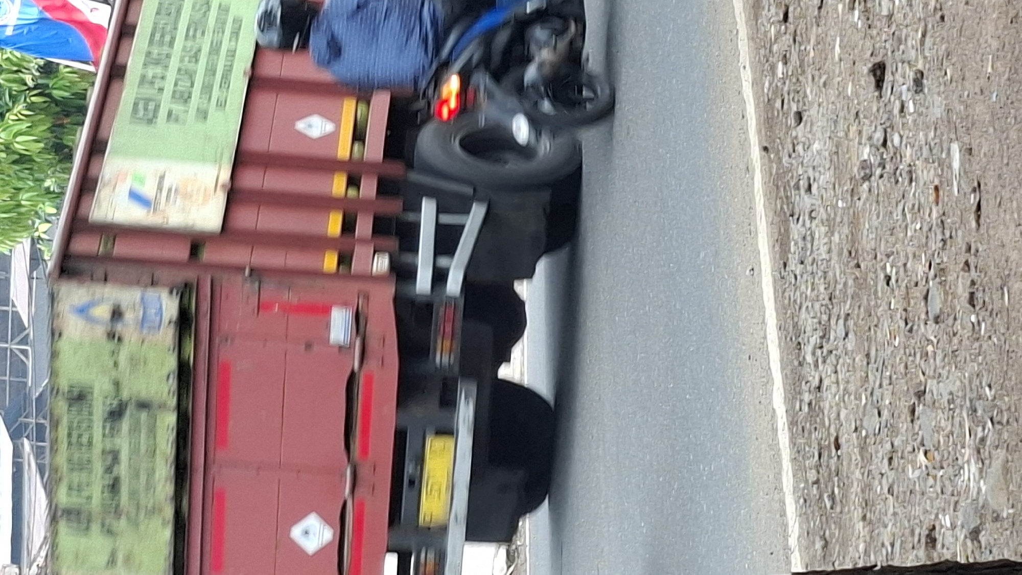 The image shows a red truck with a green cargo container in motion on a paved road. The truck has several markings and labels on its side, including a hazard symbol. A motorcyclist in a blue jacket and a black helmet is riding a motorcycle behind the truck. The environment appears to be outdoors, with some trees and foliage visible in the background. The scene is captured in motion, suggesting a street or highway setting. The location is identified as Cilaku, Indonesia.