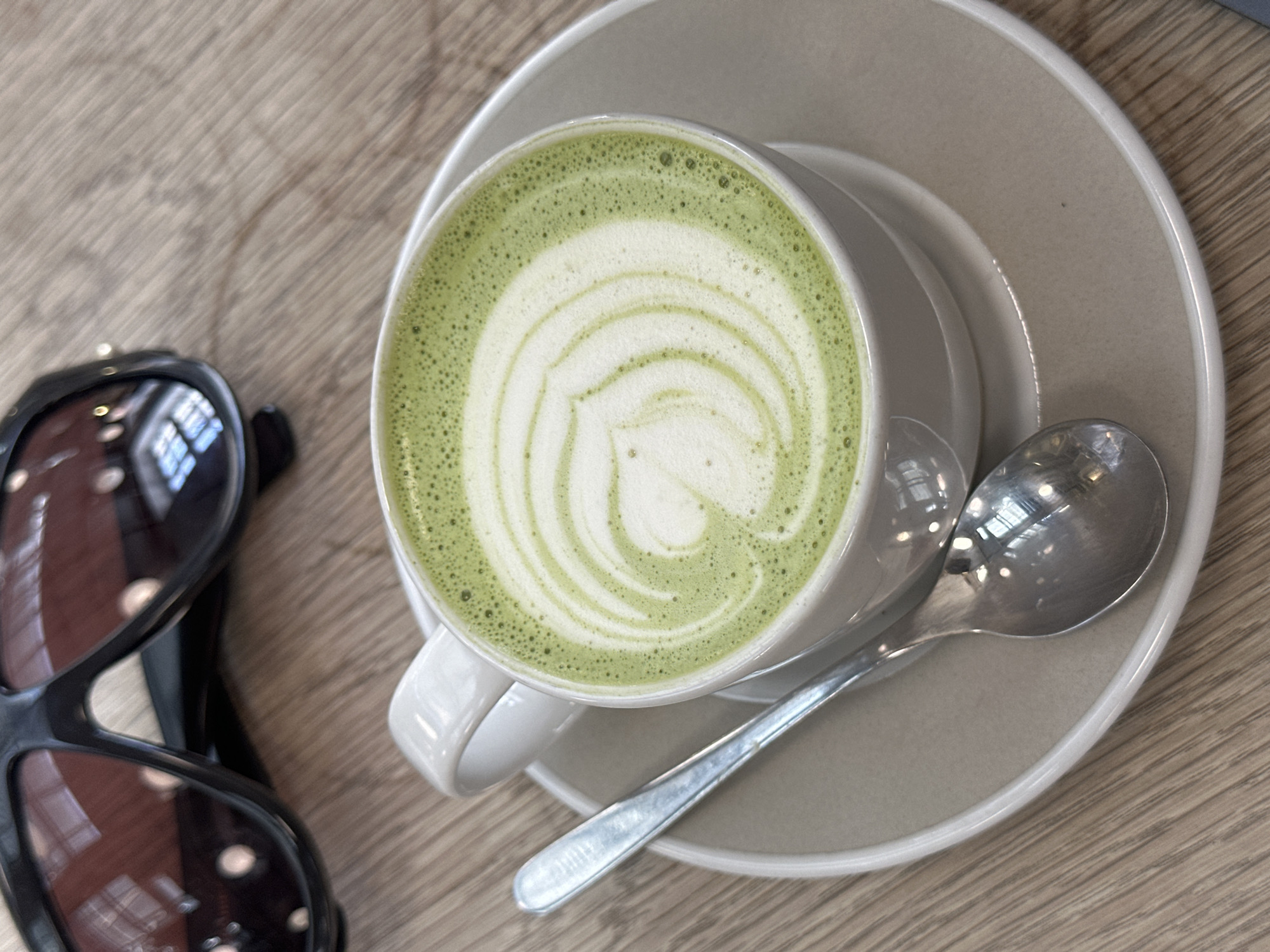 An overhead shot captures a cozy moment, featuring a matcha latte, sunglasses, and a spoon resting on a wooden table. The central subject is a white ceramic cup holding a vibrant green matcha latte, adorned with intricate white latte art forming a heart-like or spiraling pattern in its frothy surface. Small bubbles are visible on the green foam around the edge. The cup sits on a matching light grey or off-white ceramic saucer.
To the right of the cup, a silver spoon lies partially on the saucer, reflecting interior lights or window frames, suggesting an indoor setting. To the left, a pair of black-framed sunglasses rests on the wooden table. Their dark lenses also show clear reflections of what appears to be windows and the ceiling of an indoor space, further supporting a cafe or restaurant environment. The table itself is made of light-colored wood, with a distinct grain pattern visible across its surface. The overall lighting suggests it is daytime. There is no visible text in the image. The scene conveys a sense of relaxation or a break, possibly in a cafe in London, United Kingdom.