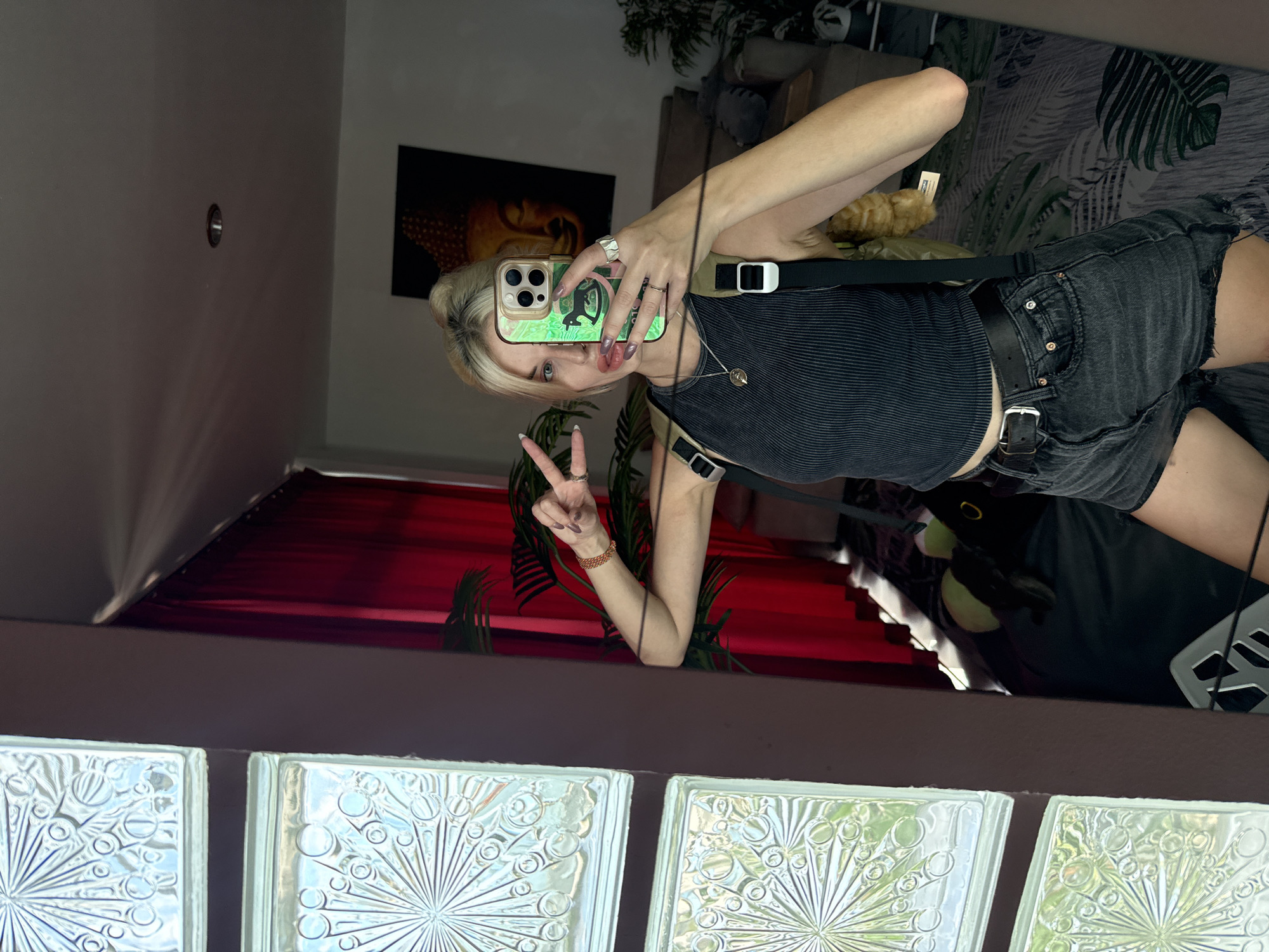 A fair-skinned person is reflected in a mirror, presumably taking a selfie. The person is wearing a black tank top, black denim shorts, and a black belt. They have blonde hair and are holding a smartphone with a green case. The person is making a peace sign with their left hand.
In the background, there is a picture of Buddha on the wall and various plants. Below the mirror, there are glass bricks with a decorative pattern.
The location is suggested to be Baan Chaweng Noi, Thailand, though this cannot be definitively confirmed from the image alone.