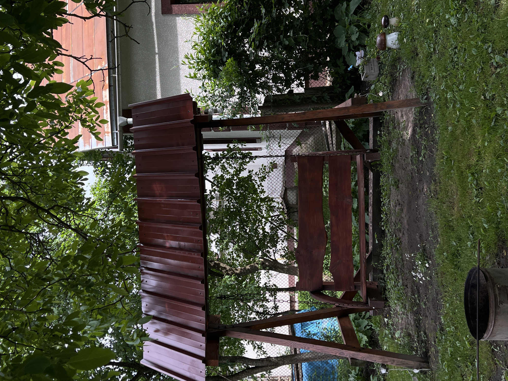 The image depicts a backyard scene in Mshanets, Ukraine, featuring a wooden garden swing with a reddish-brown corrugated metal roof. The swing structure is made of dark-stained wood, and the bench is suspended by chains.
In the foreground, the ground is a mix of sparse grass and exposed soil patches, with small white flowers visible in one dirt area. A dark, cylindrical metal object, possibly a bucket or fire pit, with a metal rod resting across its top, is positioned in the lower-left corner. Towards the right foreground, two mushroom-shaped garden ornaments and a small planter box containing a butterfly decoration are visible on the grass.
The midground is dominated by the swing set. Behind the swing on the left, a blue tarp or cover is partially visible through green foliage and a chain-link fence. Trees and dense green bushes frame the scene, with a chain-link fence running horizontally in the background, partially obscured by vegetation.
In the background, a light-colored building, likely a house, is visible. It features a reddish-orange tiled roof with a gutter and downspout system. A window with a dark frame is also discernible on the house wall. Overhead, lush green leaves from mature trees create a dense canopy, partially obscuring the sky. No people are present in the scene.