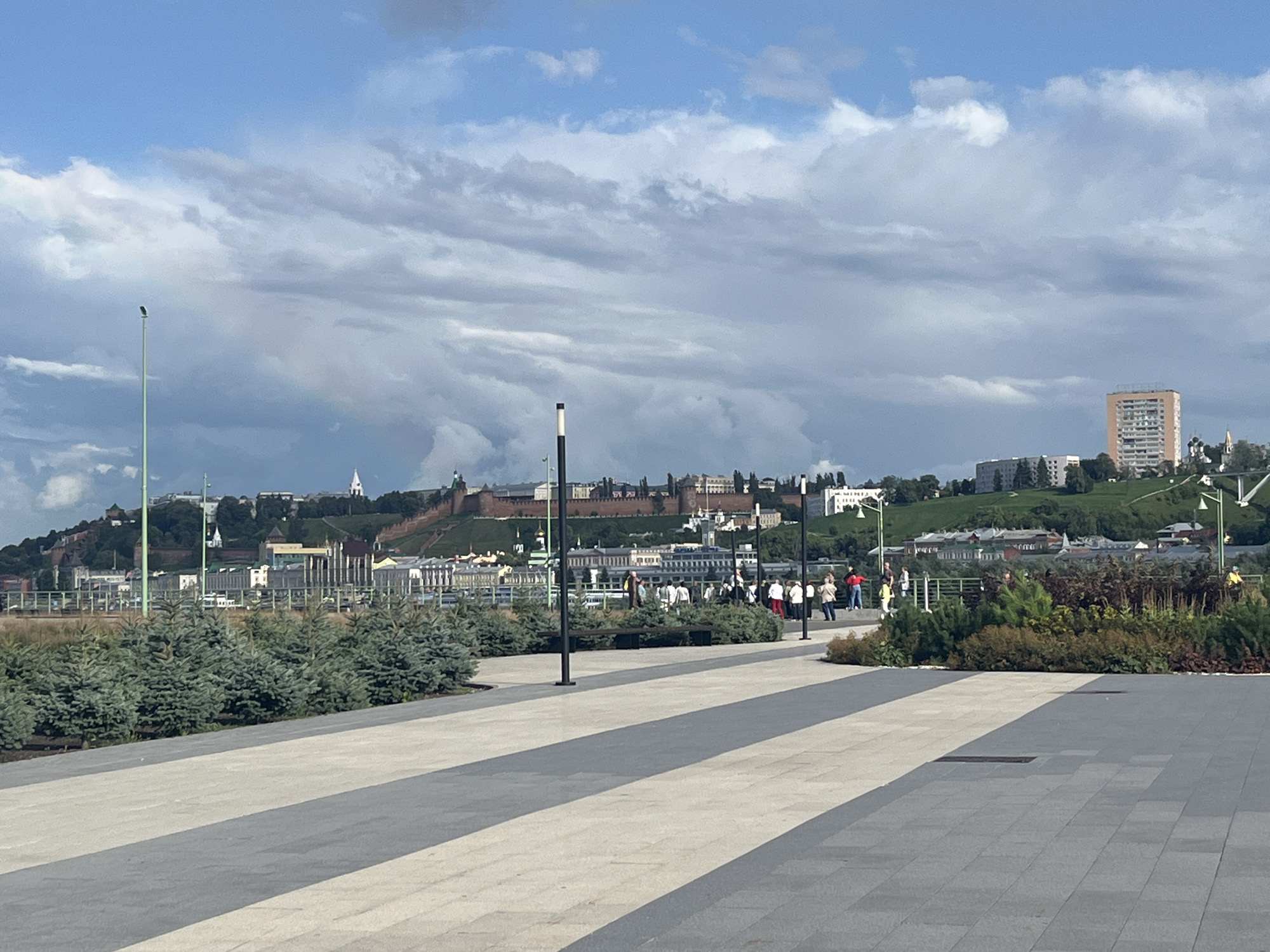 The image shows a view of the Nizhny Novgorod Kremlin and the city skyline across a plaza. In the foreground, a paved plaza with light and dark grey patterned tiles stretches towards the viewer. A row of small evergreen trees lines the edge of the plaza. In the middle ground, a group of people are walking and standing, some appearing to be tourists. Behind them, a low wall or railing separates the plaza from a sloped grassy area leading up to the Kremlin walls. The Kremlin, a historic fortress, dominates the middle and background with its red brick walls and various buildings within. Beyond the Kremlin, the city of Nizhny Novgorod unfolds, with numerous buildings, including a tall, orange apartment building on the right. The sky above is partly cloudy with patches of blue sky. A faint rainbow is visible in the sky to the left of the Kremlin. The scene appears to be a public space on a pleasant day.