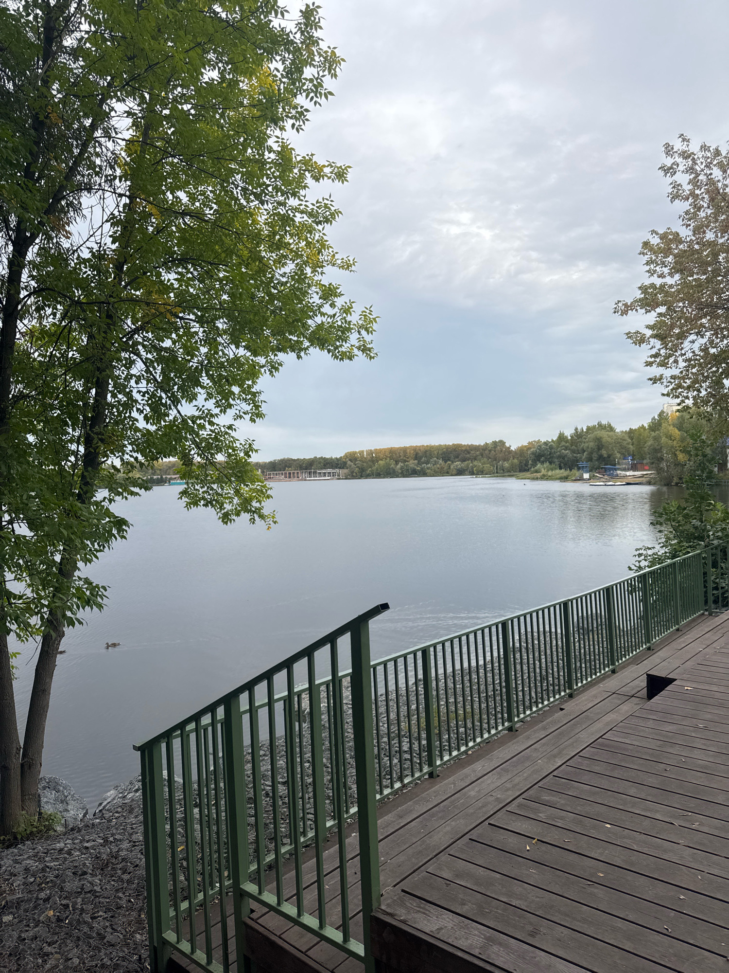 The image depicts a serene lake scene, likely in Ufa, Russia, given the provided context of the city. In the foreground, a wooden deck leads to a green metal railing overlooking the water. The railing follows a set of stairs descending towards the rocky shoreline. The lake is calm, with a few ducks visible swimming. On the opposite bank, a dense line of trees forms the horizon, with distant structures suggesting a park or recreational area. The sky is overcast, with soft, diffused light, indicating a cloudy day. The overall atmosphere is peaceful and natural.