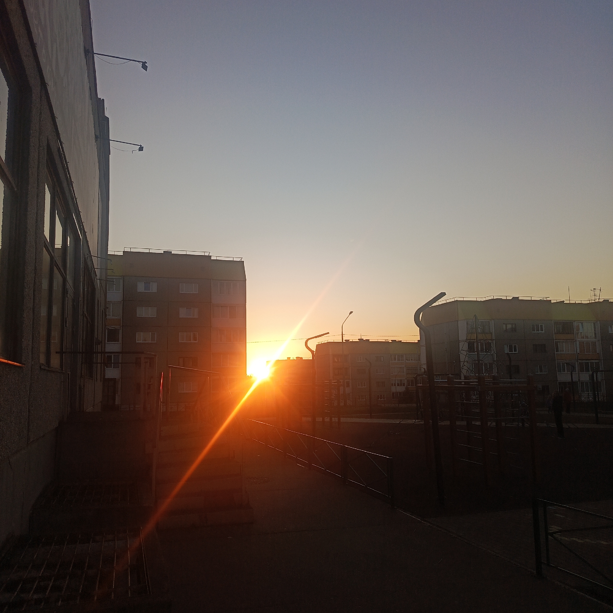 The image captures a sunset in what appears to be a residential area of Марковский, Russia. The sun, partially obscured by apartment buildings, casts a strong orange glow and lens flare. In the foreground, a school building with large windows is visible on the left. A fenced playground area is in the center, with various play structures. A figure, possibly a person, is silhouetted near the right side of the frame. The sky transitions from a light orange near the horizon to a pale blue above. Streetlights are visible between the buildings. The overall scene is urban, with the focus on the sunset over the built environment.