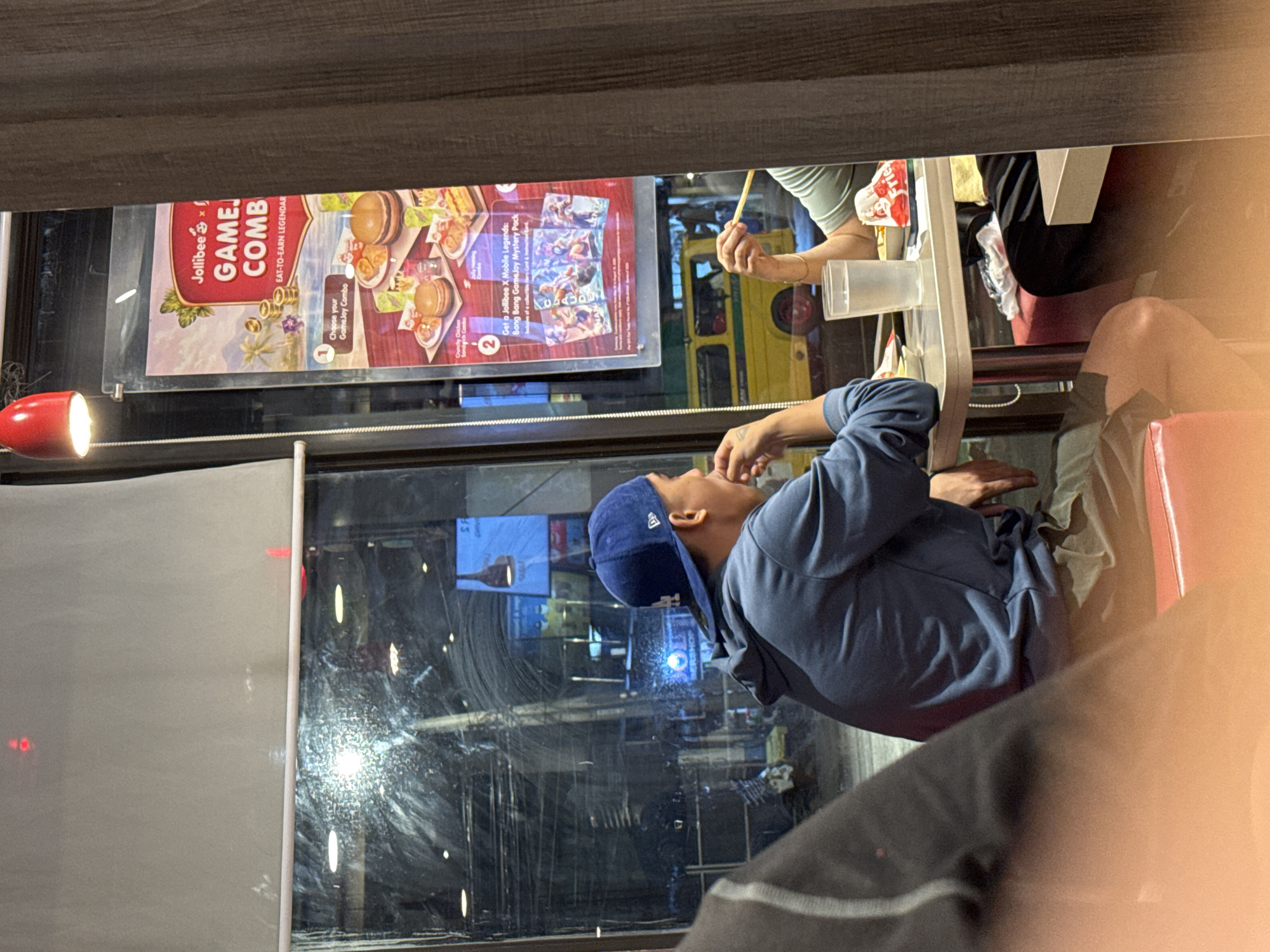 The image captures a candid moment inside a Jollibee restaurant in Davao City, Philippines, likely during the evening or night. The primary subject is a man wearing a blue hoodie and a blue baseball cap, seen in profile, looking upwards at a Jollibee menu board displayed behind a large window. His right hand is near his mouth.
Reflected in the window are glimpses of the city outside, including what appears to be a yellow jeepney or similar public transport vehicle, along with various other lights and structures. A red lamp casts a glow near the window on the left.
On the right side of the frame, parts of another person are visible, specifically an arm and hand holding chopsticks over a table, suggesting they are eating or about to eat. A plastic cup is also on the table.
The prominent Jollibee menu board advertises a "Jollibee x GAME COMB" with the tagline "EAT-TO-EARN LEGENDAR". It features images of burgers and states, "1 Choose your Game.Joy Combo" and "2 Get a Jollibee X Mobile Legends: Bang Bang Game.Joy Mystery Pack". Text "CLAUDE" and "Fries" are also visible on promotional items. The scene is illuminated by interior lights, contrasting with the dark reflections outside.