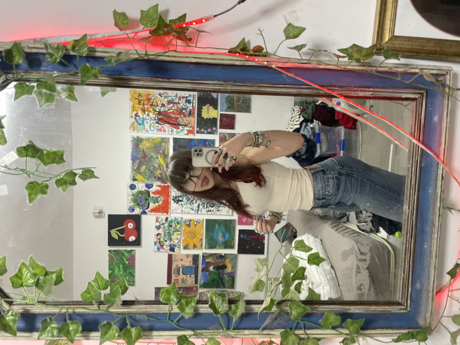 A person with bangs, glasses, a light-colored top, and jeans takes a mirror selfie. The mirror has a blue frame, and ivy and red string lights are wrapped around it. The wall in the background has numerous small artworks displayed, creating a colorful and eclectic aesthetic. There's also a chair and some clothes visible behind the subject. The image appears to be rotated.