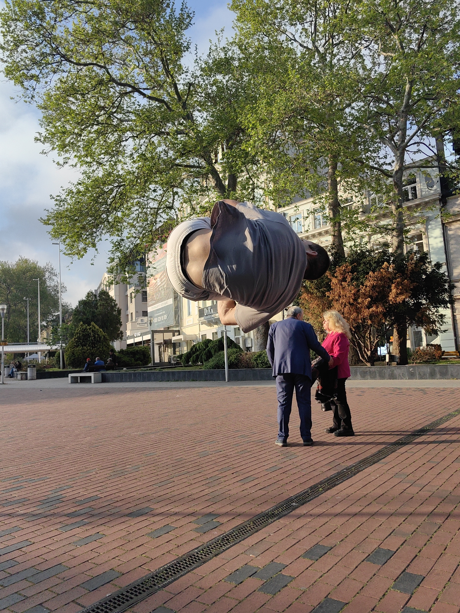 A man is performing a handstand on a public square in Unknown, Unknown, with a couple standing to his right, appearing to watch him. The square is paved with red bricks, and a drainage grate runs along the lower right. Behind the couple is a raised garden bed with bushes and small trees. Further behind, buildings line the square, with trees in the foreground. The sky is partially cloudy.