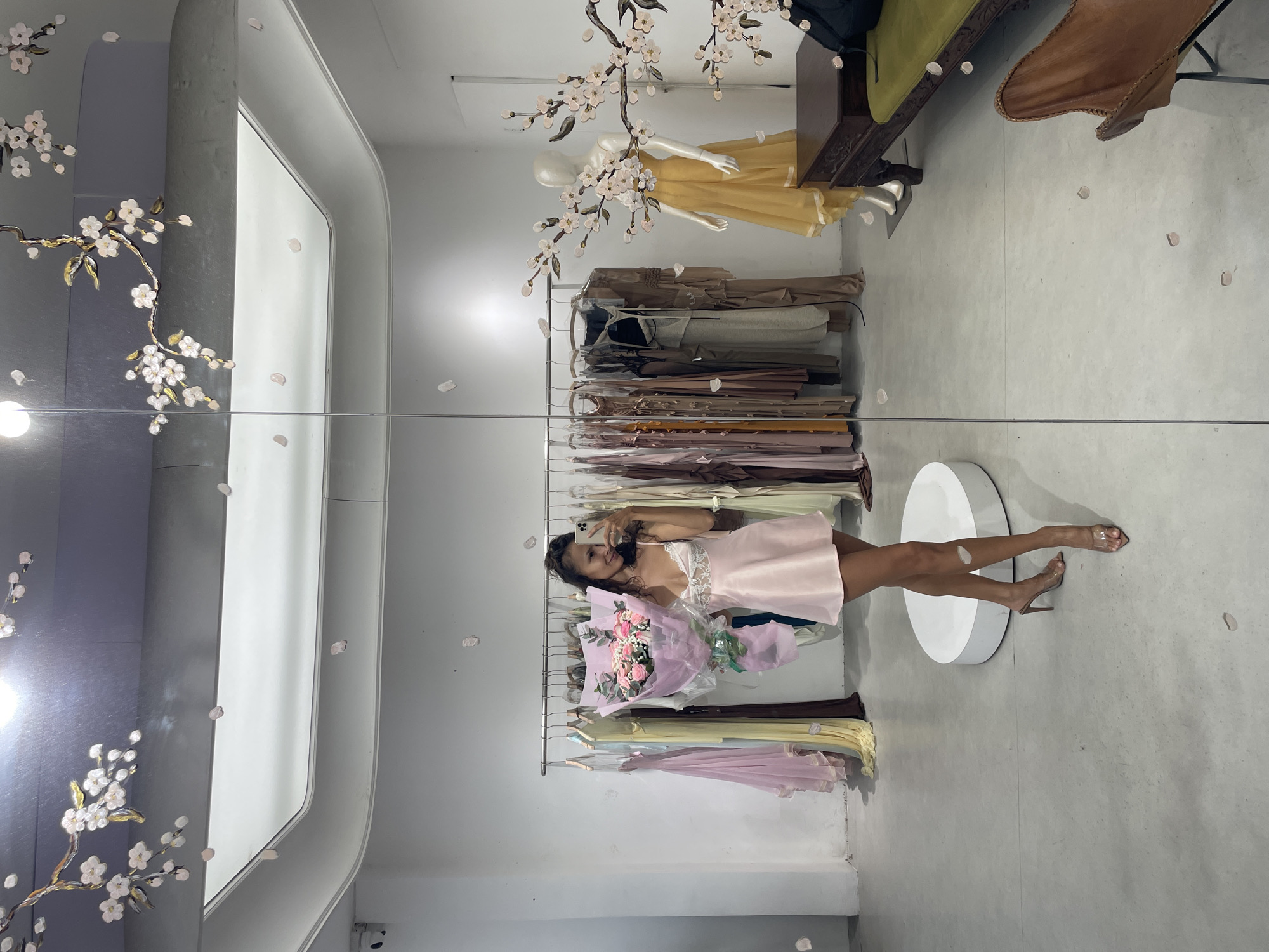 This image depicts a mirror selfie taken by a woman inside what appears to be a retail boutique or showroom. The location in the city of Thủ Đức, Vietnam, cannot be definitively ascertained from the visual content of the image.
In the center of the image, reflected in a large, floor-to-ceiling mirror, a female subject is visible from mid-thigh up. She possesses dark, wavy hair, and is smiling slightly while looking towards her smartphone, which she holds in her left hand, positioned for a selfie. Her right arm cradles a large bouquet of approximately two dozen pink roses, wrapped in clear plastic film and translucent pink paper, secured with a green ribbon at the base. She is wearing a short, light pink satin slip dress featuring white lace detailing on the bust. Her footwear consists of clear high-heeled mules. A delicate necklace is visible around her neck. She stands on a light grey concrete or polished cement floor, with a circular white display platform visible beneath her feet.
The background, largely reflected in the mirror, shows the interior of the establishment. To the left of the subject, a clothing rack displays several long, flowing dresses in pastel shades, including light pink, yellow, and possibly blue or cream. To her right, another clothing rack holds dresses predominantly in earthy tones, such as various shades of brown, beige, and grey. Further to the right in the reflection, an armless white mannequin wears a long, draped mustard-yellow dress. Below the mannequin, a dark brown, carved wooden console table or bench is visible, with a green upholstered cushion on its surface. In the bottom right corner of the reflected space, a portion of a brown leather butterfly chair is discernible.
The walls are painted white, and the floor is a uniform light grey. The ceiling is white, with prominent light fixtures and a dark grey air duct or structural element running across it. Decorative elements include artificial cherry blossom branches with white flowers and golden leaves, strategically placed on the ceiling, upper walls, and possibly directly on the mirror surface. Numerous light pink petals are scattered across the floor and adhered to the mirror, contributing to a decorative "falling petal" effect. The space is brightly illuminated by overhead lighting.