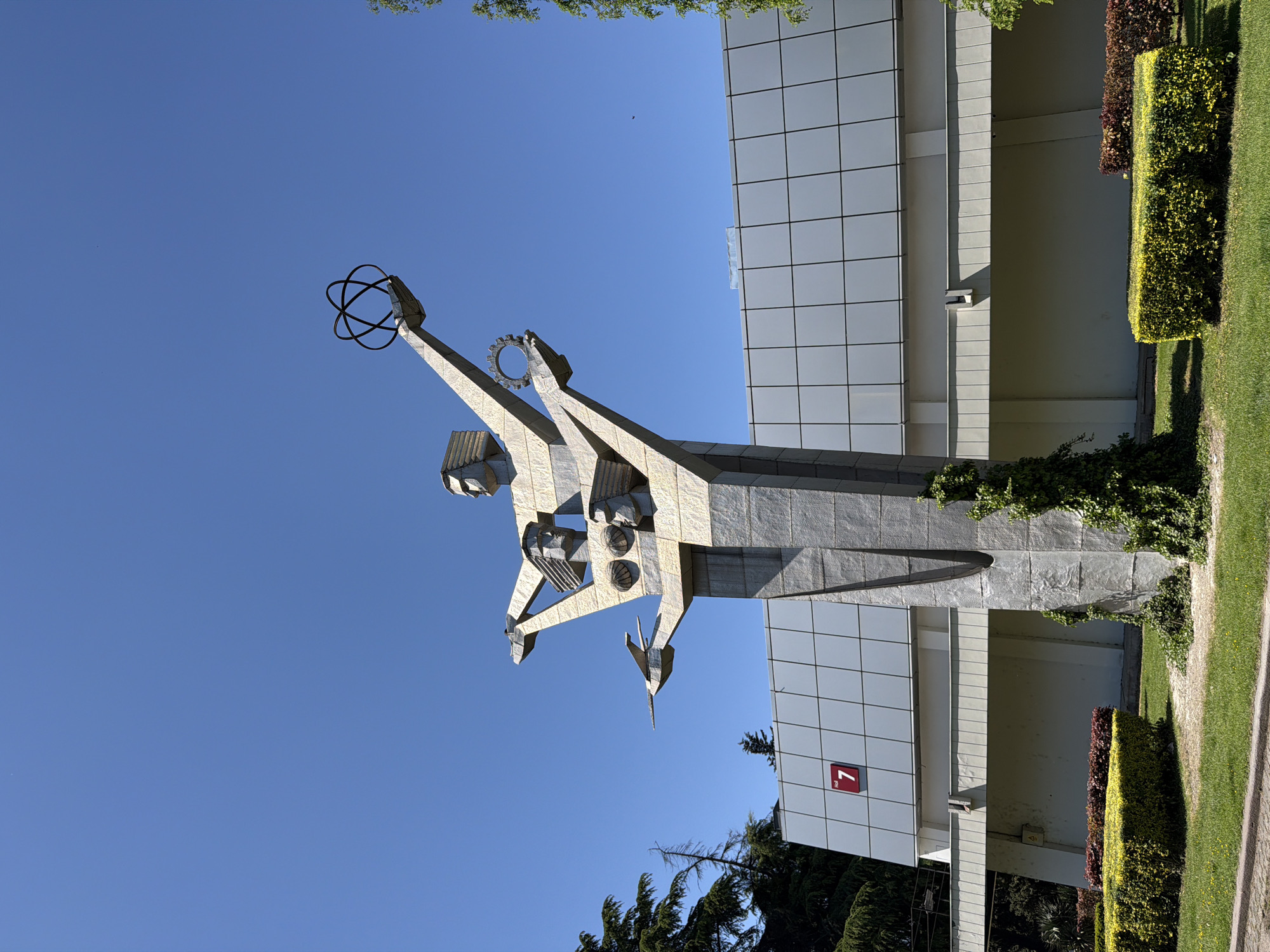 A low-angle, outdoor photograph shows a monument and building against a bright blue sky. The monument is constructed from gray, textured material. It features two stylized figures reaching upwards, one holding a circular object with interconnected rings, and the other holding a gear-like ring. A small, stylized object resembling a bird is also part of the sculpture. The building behind the monument has a facade of square, white panels. A red square containing a white number 7 is visible on the building’s lower portion. Green shrubbery and some trees are present at the base of the building and around the monument. This is the “History of Georgian Medicine” monument, located near Tbilisi State Medical University in Tbilisi, Georgia.