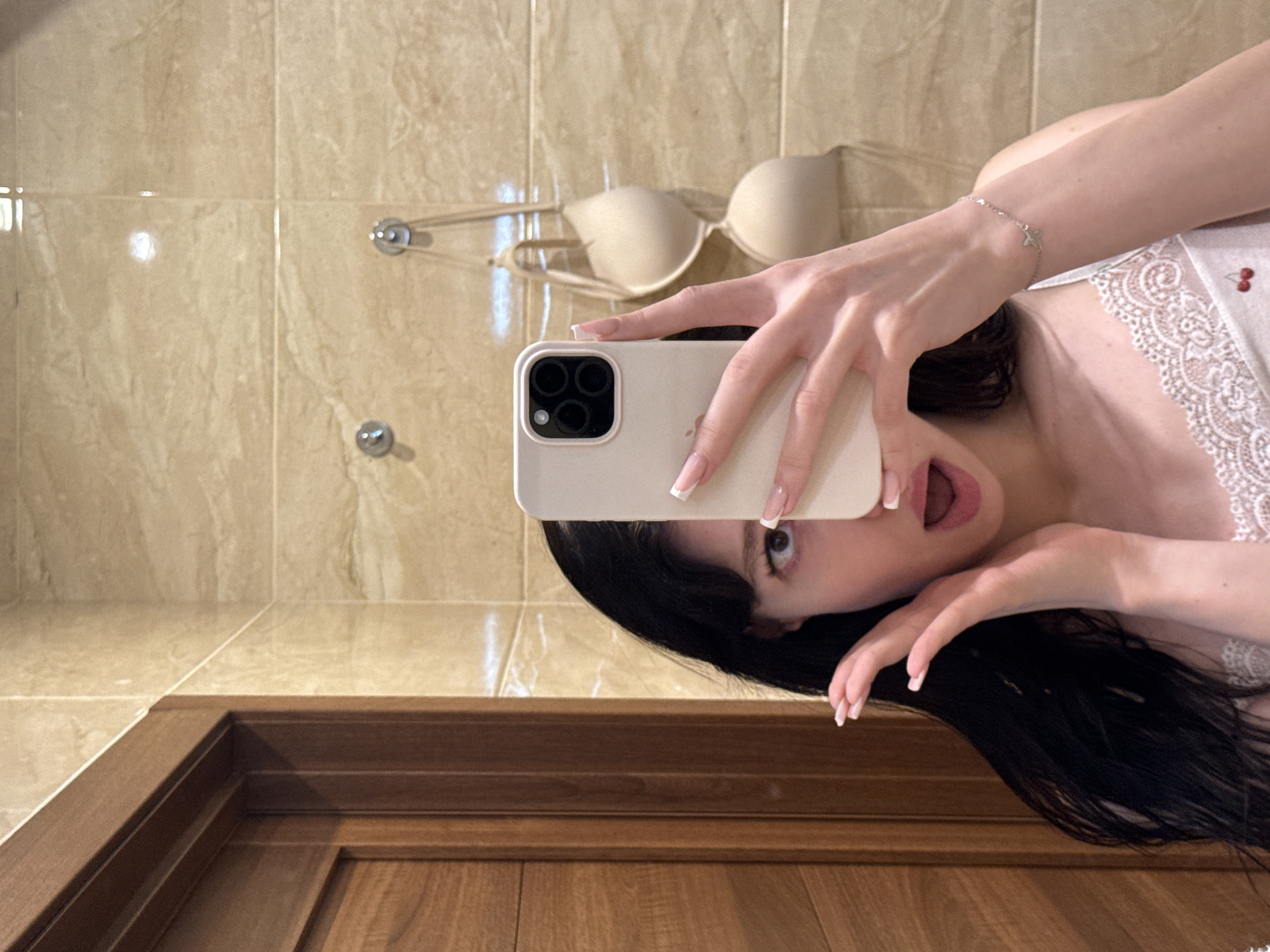 A young woman with long, dark hair is taking a selfie in a bathroom. She is wearing a white camisole with lace trim and a cherry print. She is holding a white smartphone in her right hand, which is angled to capture her reflection in a mirror. Her left hand is raised to her face, with her fingers spread. Her mouth is open in a surprised or playful expression, and her eyes are wide. In the background, a beige tiled wall is visible, with a wooden door on the left. A beige bra is hanging on hooks on the wall behind her. The lighting is bright and even, suggesting an indoor setting. The overall impression is casual and intimate, possibly a candid moment captured at home.