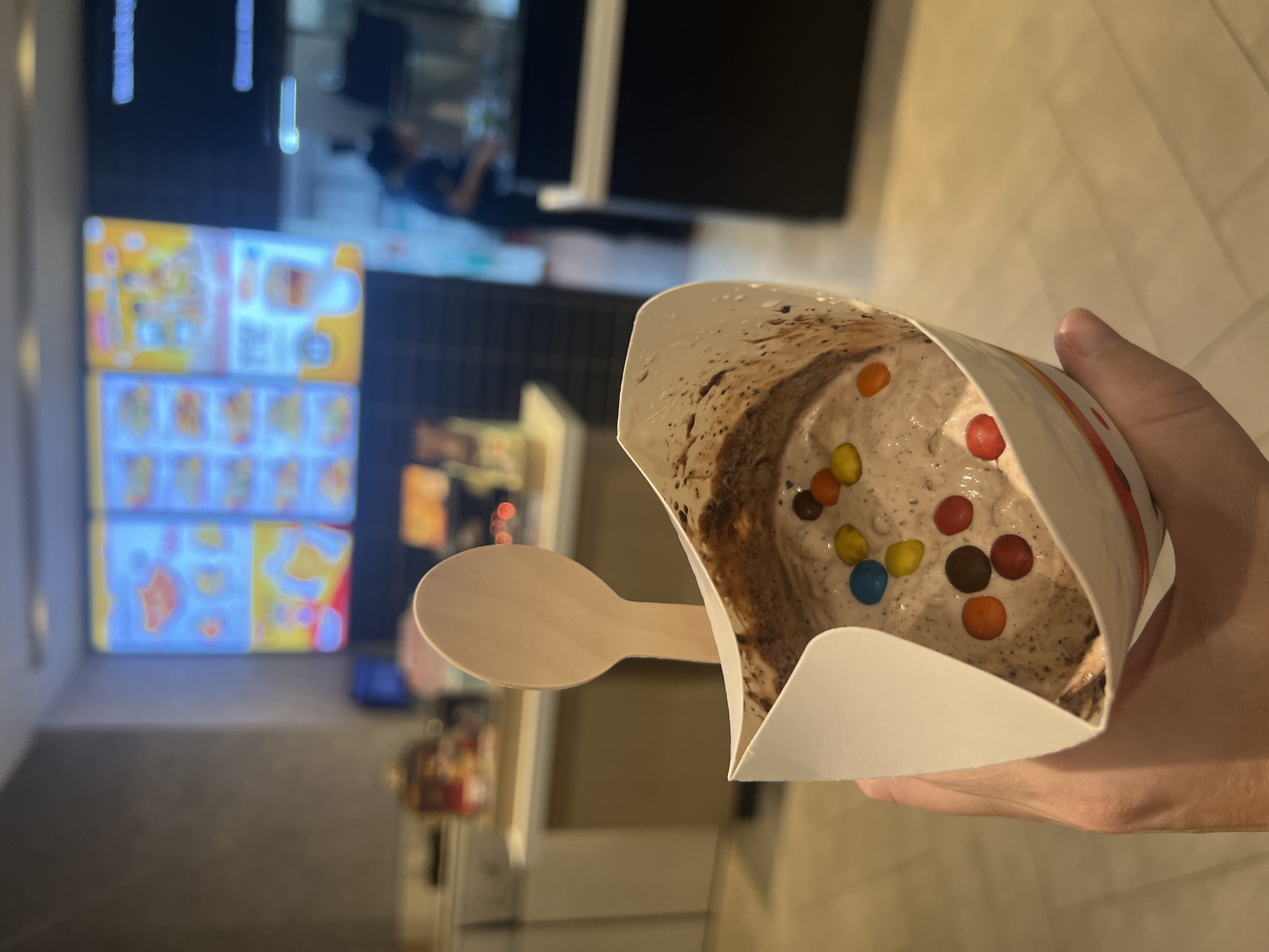 A person's hand holds a white paper cup filled with a brown, creamy substance, likely ice cream, which is topped with several colorful candy-coated chocolates. A wooden spoon is inserted into the cup. In the background, out of focus, is the interior of what appears to be a fast-food establishment with a digital menu board displaying colorful images of food items. Another person, a staff member, is visible in the background, seemingly working behind a counter. The environment suggests an indoor setting with artificial lighting and a tiled floor.