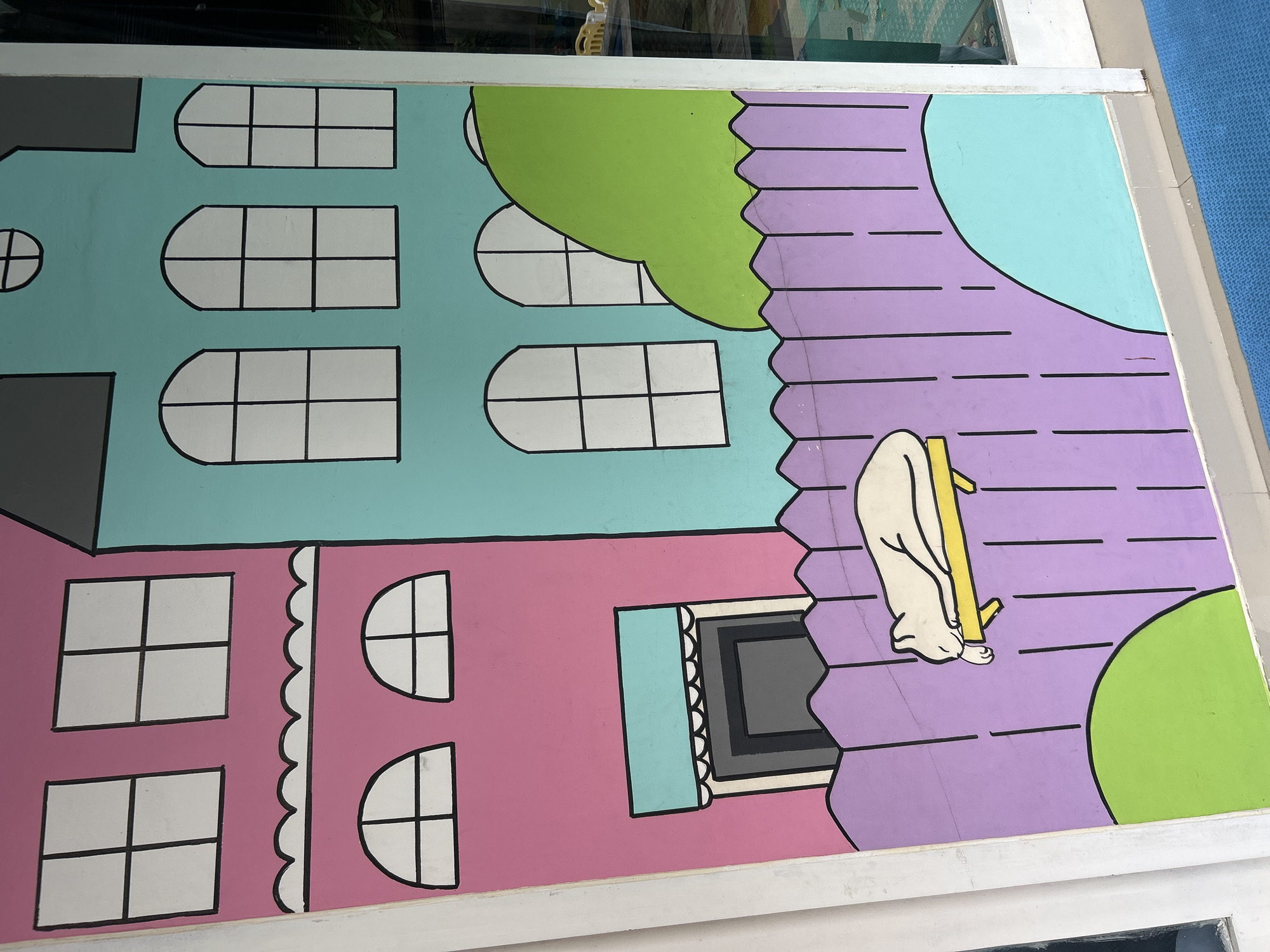 The image is a close-up of a mural painted on an outdoor wall, likely in Kuta, Indonesia. The mural depicts a stylized cartoon scene with two houses side-by-side. The left house is painted pink and features two square windows on the upper level and two arched windows on the lower level. Below these windows is a doorway with a teal awning and scalloped trim. The right house is painted teal and has three arched windows on the upper level, a small circular window above, and another arched window below. A large green, rounded shape, possibly representing a bush or hill, is visible behind the teal house. In front of the houses, a purple picket fence runs across the lower portion of the mural. A white cartoon cat is depicted sleeping on a yellow bench in front of the fence. The mural is rendered in bold, simple lines and bright, flat colors. The wall appears to be part of an exterior building, possibly a shop or cafe, with a white frame on the right side of the image suggesting an adjacent doorway or window.