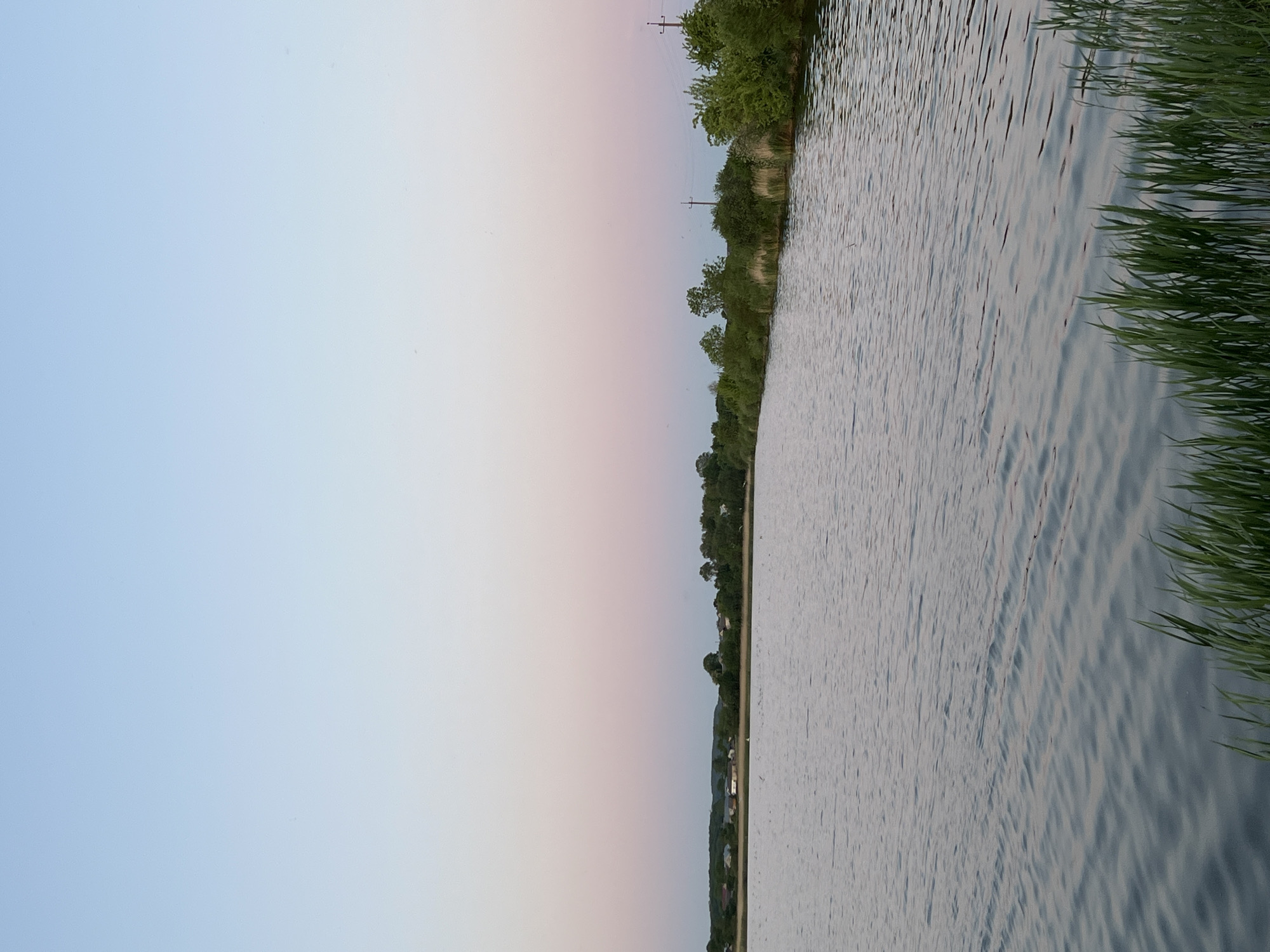 The image shows a landscape scene, possibly taken in Reniv, Ukraine. The right side of the image is dominated by water, likely a river or lake, with some visible wave patterns on its surface. Reeds grow along the water's edge. The left side is a gradient of sky, transitioning from a light blue near the top to pinkish hues near the horizon. A line of trees and vegetation separates the sky from the water. In the distance, hints of buildings and structures are visible along the shore opposite the viewer. Overhead power lines are also present.