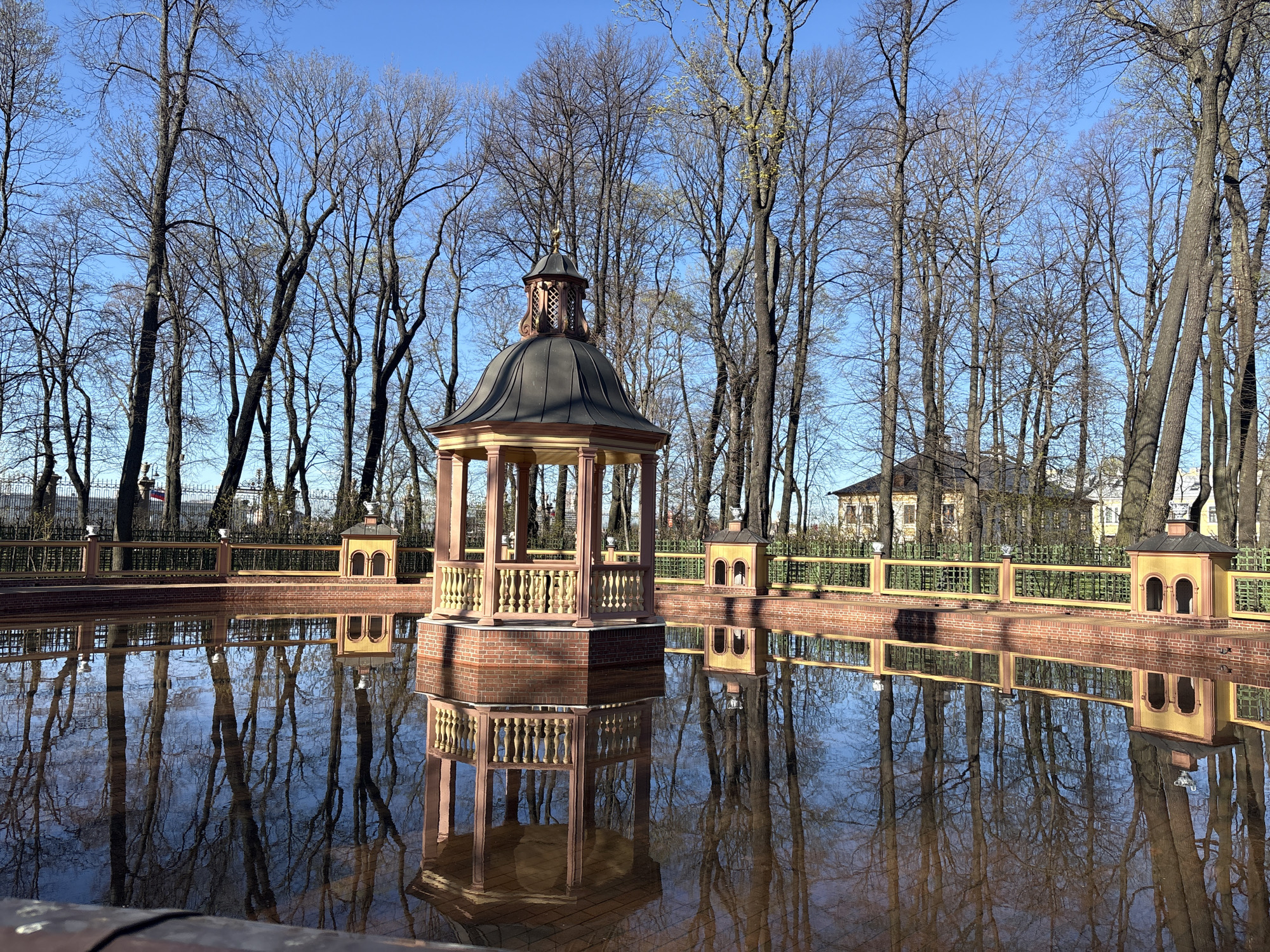 The image depicts a decorative pavilion situated within a pond or reflective pool. The pavilion has a dark, rounded roof and is supported by columns with railings. It appears to be constructed of brick and painted in light tan and brown shades. The pond reflects the pavilion and surrounding trees, creating a mirrored effect. The trees are tall and bare, suggesting either late autumn or early spring. A brick border surrounds the pond, with smaller, decorative structures at regular intervals. In the background, a light-colored building can be seen through the trees. The sky is clear and blue. This appears to be the Chinese Arbor in the Alexander Garden, Saint Petersburg, Russia.