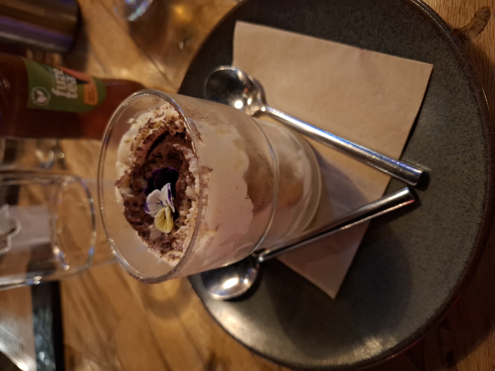 A close-up, eye-level shot shows a dessert in a clear glass, topped with whipped cream, cocoa powder, and two small edible flowers. The glass sits on a brown paper napkin, which is placed on a dark, textured plate. Two silver spoons are positioned on either side of the glass, with their handles extending outward. In the background, a blurred glass of water and a bottle of "Fuze Tea" are visible on a wooden table. The overall lighting is dim and warm.