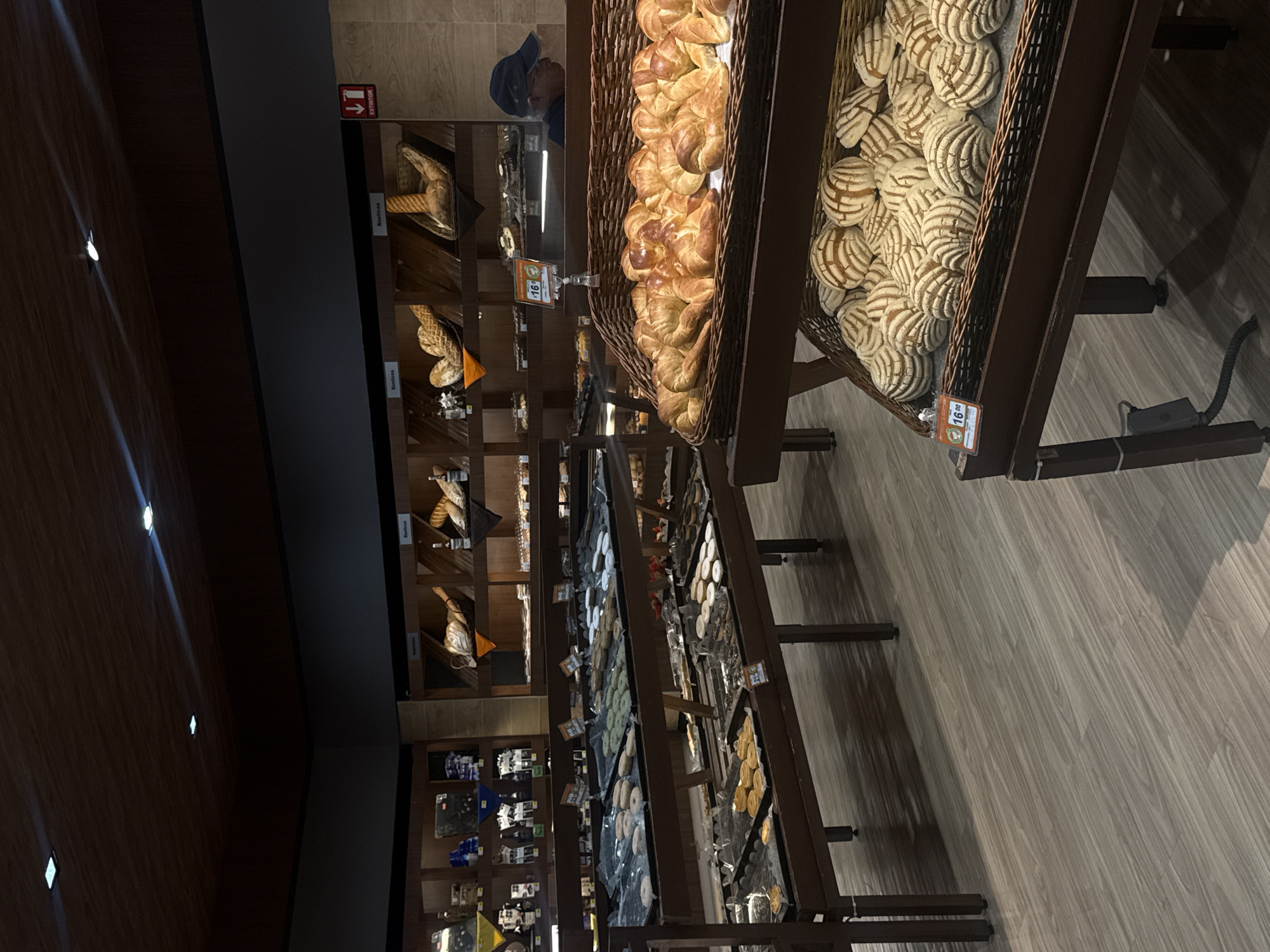 The image shows an interior view of a bakery, with a variety of baked goods on display. At the forefront are large baskets filled with pastries, including croissants and conchas (a type of Mexican sweet bread). Shelves in the background hold various types of bread and other baked items, with price tags visible near some of the goods. A person wearing a blue cap is partially visible in the background behind the pastries. The bakery has a modern, well-lit environment with wooden shelves and flooring.