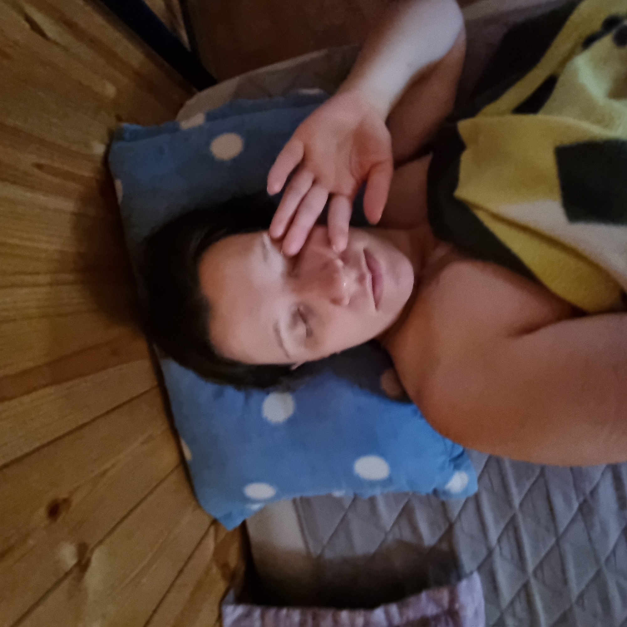 A person lies in bed, with their right hand raised to their closed right eye, as if waking up. The person is partially covered by a blanket with a yellow and black geometric pattern. The person is on a bed with a blue and white polka-dot pillow and a quilted grey mattress. The background consists of wooden paneling. The image appears to have been taken in Dunnae-myeon, South Korea.
