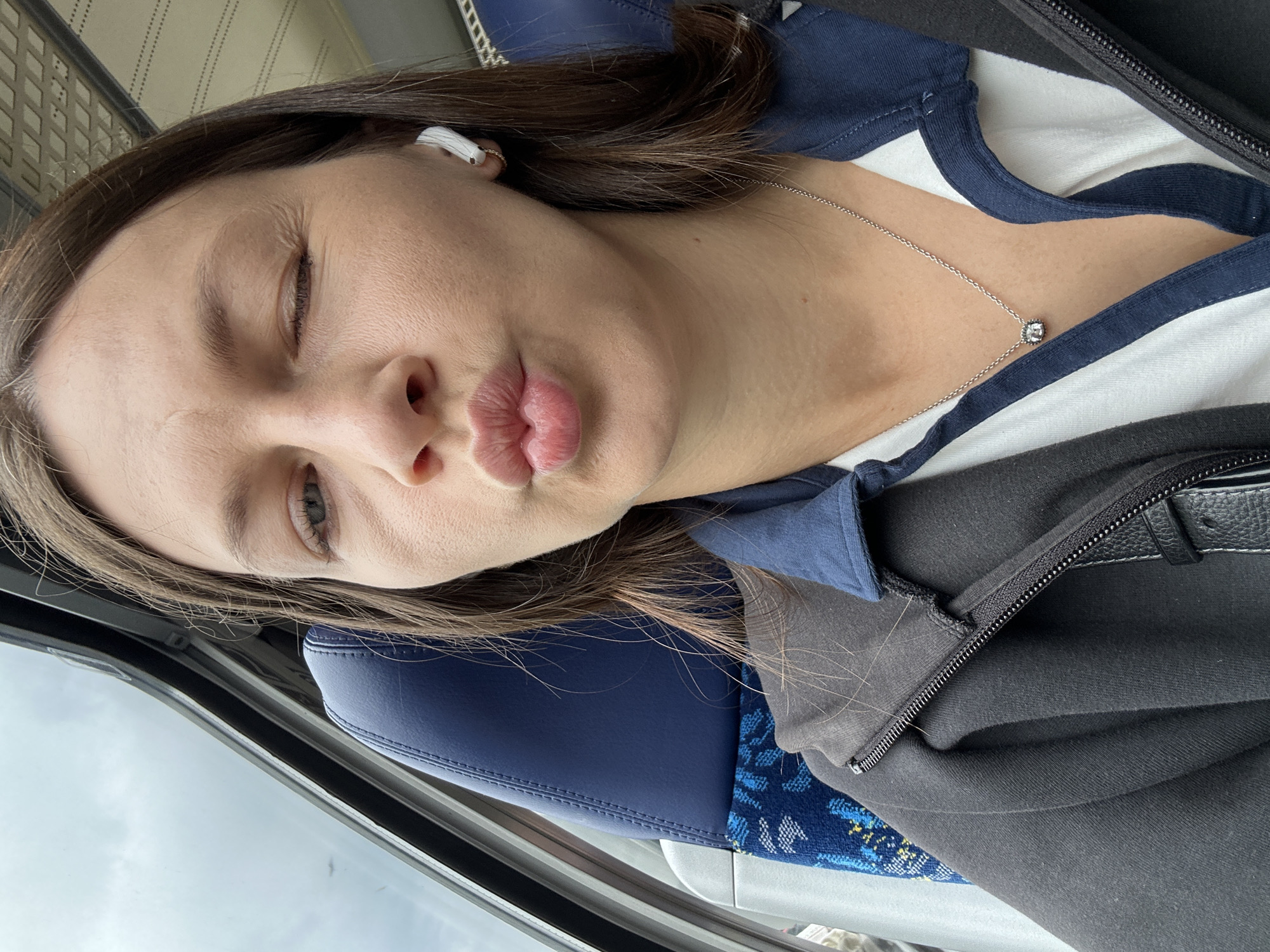 A young woman, likely in Buxtehude, Germany, is captured in a tilted, close-up selfie, making a playful "kiss" face with pursed lips. Her right eye is closed, while her left eye is slightly open. She has long, dark brown hair and is wearing a white AirPod in her right ear.
She is dressed in a white top with a navy blue collar and stripes, layered under a black zippered jacket or hoodie. A delicate silver chain necklace with a small, square, clear pendant rests on her chest. The setting appears to be a public transport vehicle, indicated by the distinctive blue and patterned fabric of the seat and the view of a cloudy sky through the window. The light suggests it is daytime. There is no visible text in the image.