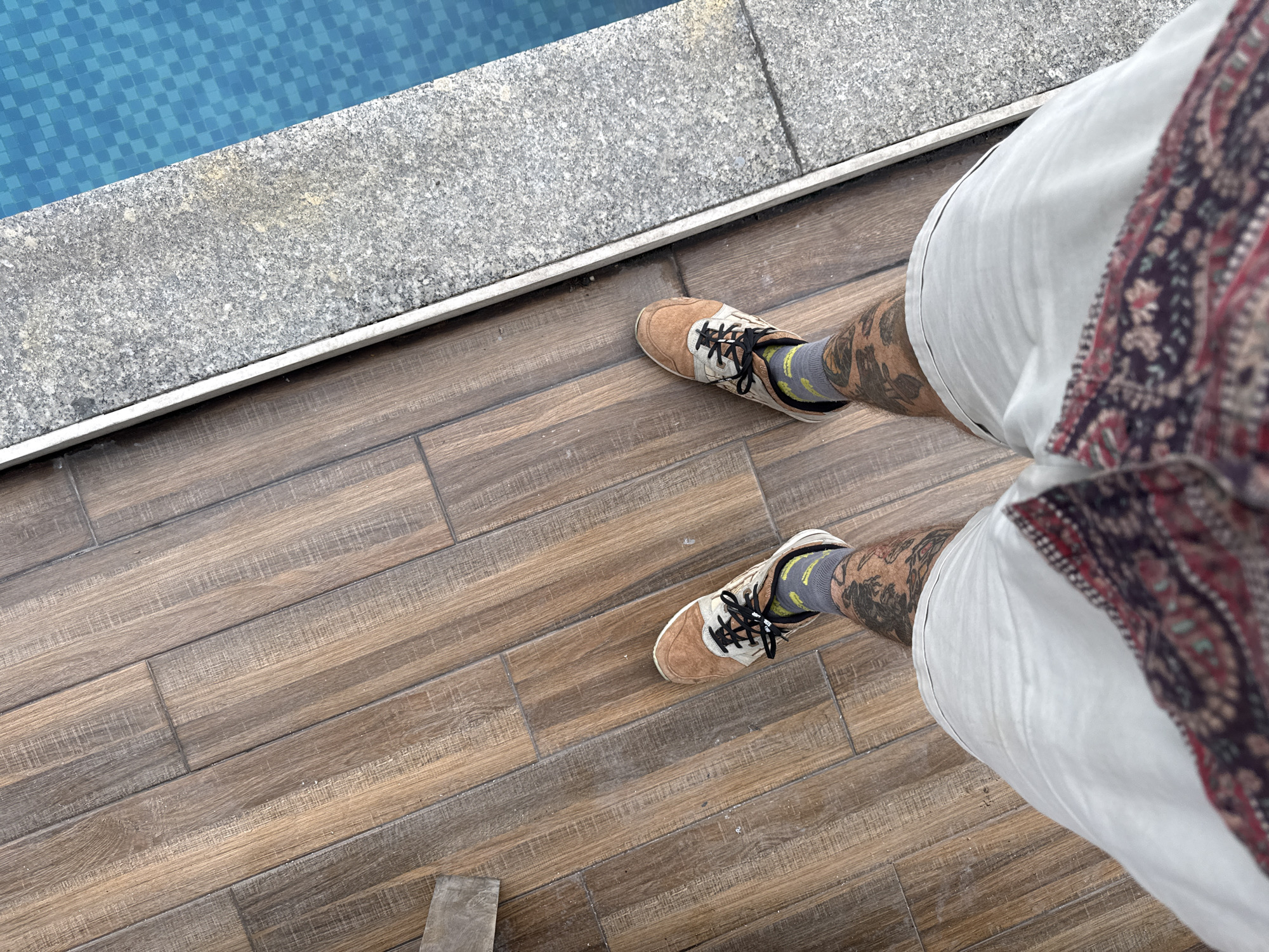 A first-person, high-angle shot shows a person standing on a tiled wooden floor near a pool, likely in Đà Nẵng, Vietnam. The pool's edge is visible at the top left corner, marked by light blue tiles and a gray stone border. The person is wearing tan and white sneakers with black laces and gray socks with a yellow pattern. Their legs are visible below white shorts, with tattoo art on the lower legs. Part of a patterned shirt is seen on the right side of the frame.