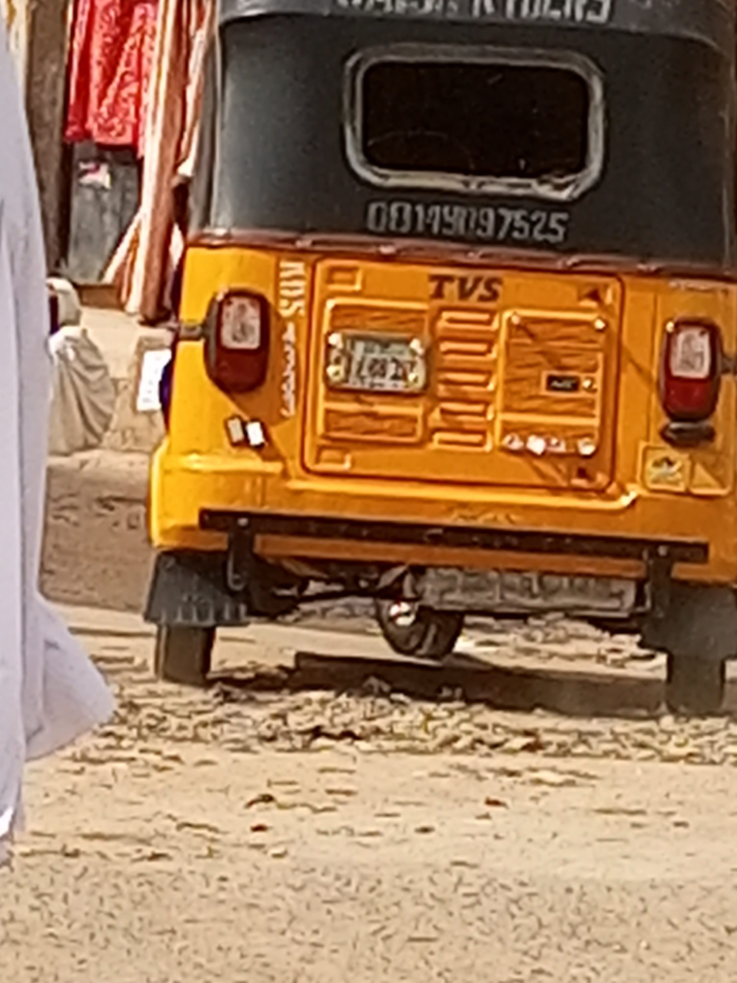 The image shows the rear of a yellow TVS tricycle parked on a dusty surface. The tricycle has a black roof with the text "KDS RIDERS" at the top and the number "08149097525" below the rear window. The license plate is partially visible. To the left, a person in white clothing is partially visible in the foreground. In the background, fabric is draped over an unidentified structure. The location is identified as Maiburiji, Nigeria.