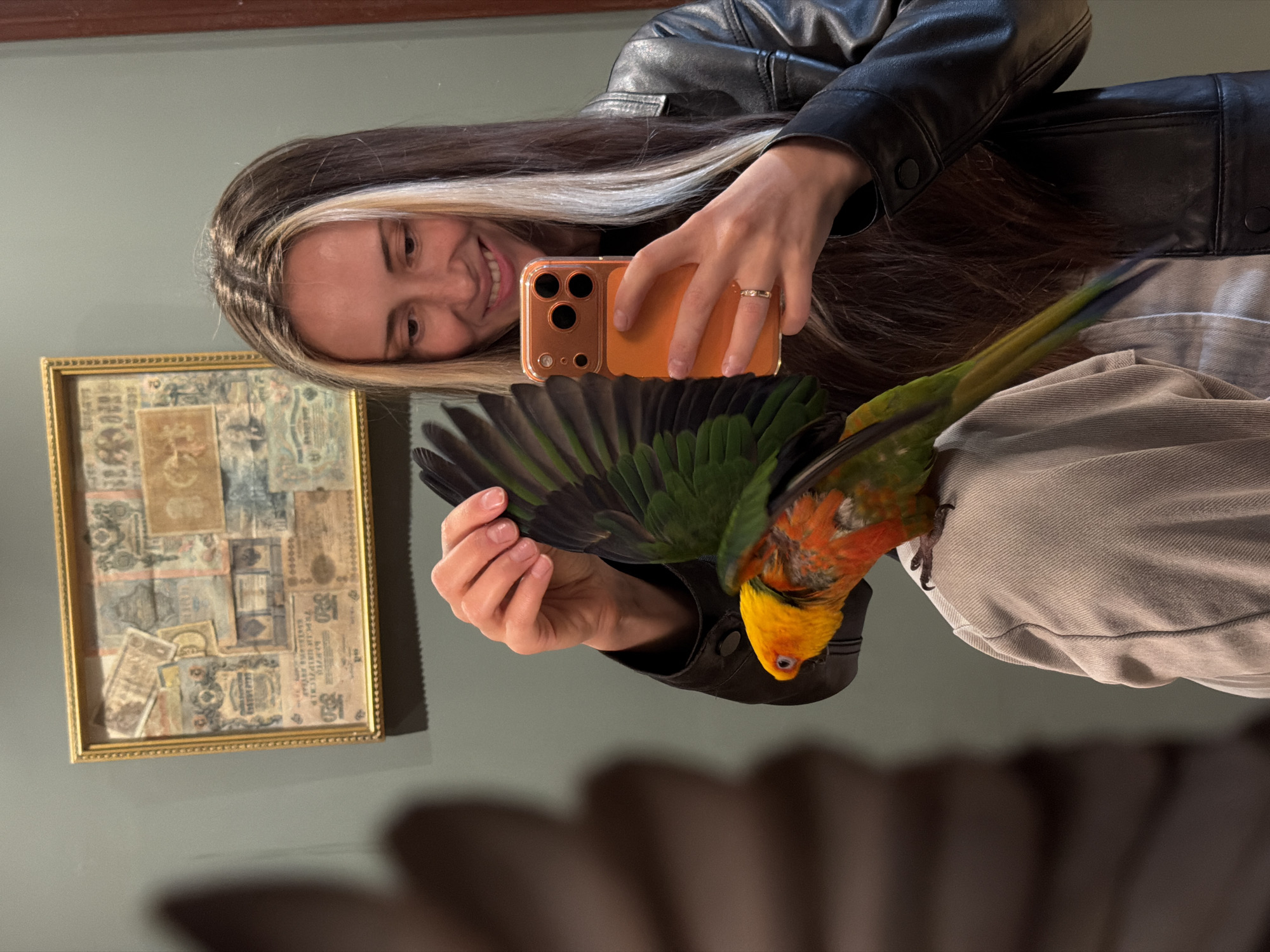 A young woman is taking a mirror selfie in what appears to be a home. She is wearing a black leather jacket and has long, dark hair with blonde highlights. Her face is visible in the reflection, and she is smiling. She is holding an orange smartphone to take the picture. A small parrot with green, orange, and yellow feathers is perched on her shoulder, with its wings partially spread. In the background, to the left, there is a framed picture containing various old stamps or postcards. The wall behind her is painted a muted green. The overall lighting suggests it is daytime, possibly indoors with artificial lighting.