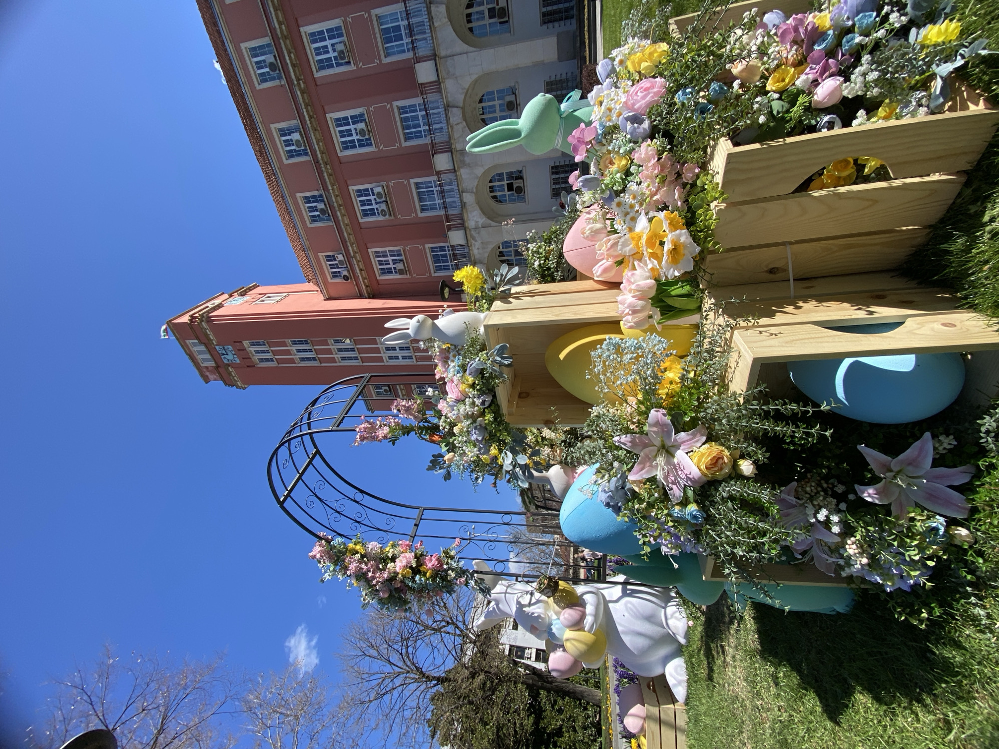 The image shows an Easter decoration in Pleven, Bulgaria. The decoration includes wooden crates arranged in a tiered fashion, filled with colorful artificial flowers and Easter eggs. One crate features a pink egg and a green bunny. A white ceramic bunny is perched on top of a crate. An arch decorated with flowers is also part of the display. In the background, a pink building with many windows can be seen under a clear blue sky. The decoration is set on a patch of green grass, and bare trees are visible to the left.